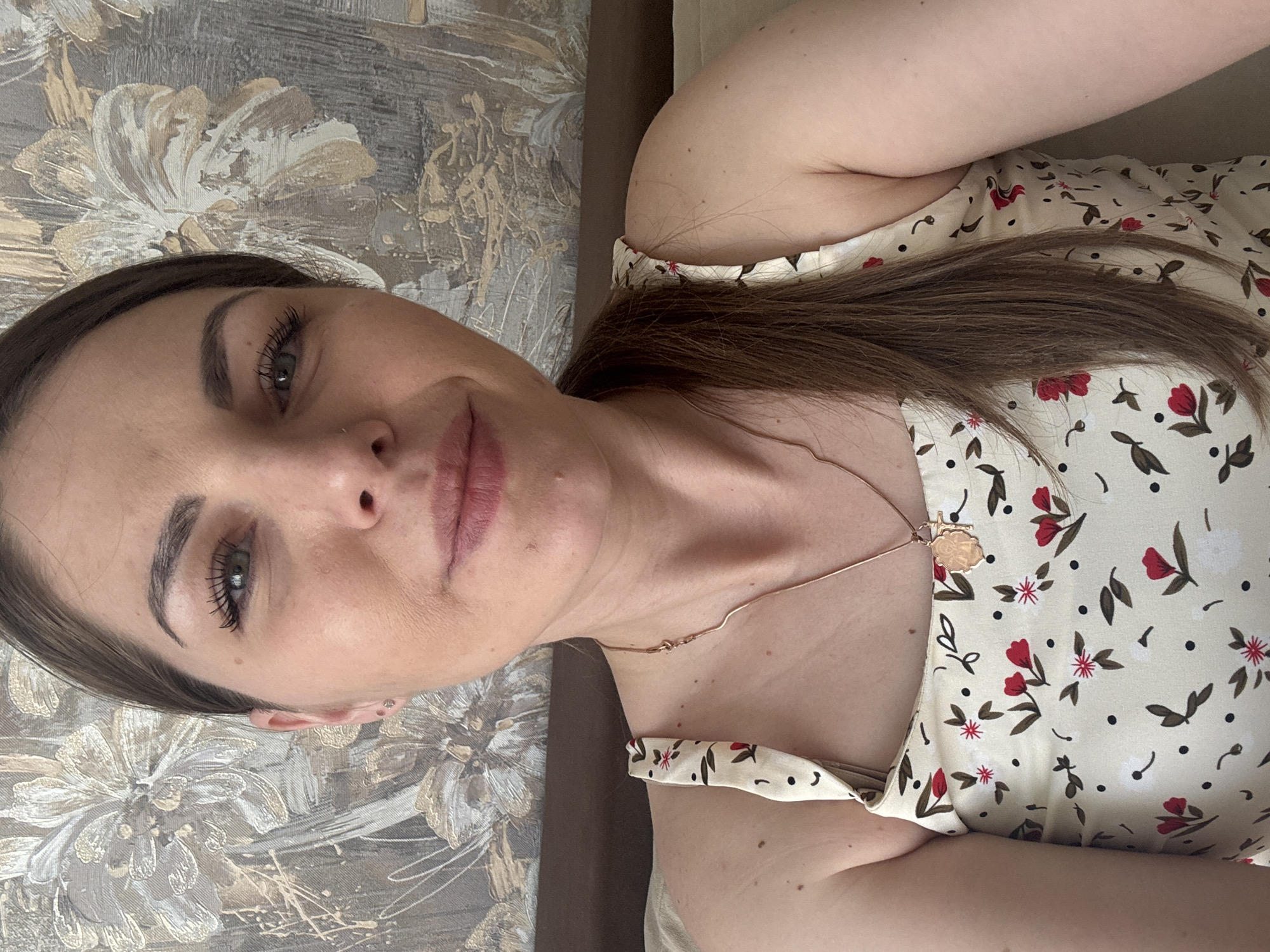 A woman is pictured in a close-up selfie. She has brown hair pulled back in a ponytail, fair skin, and visible eyelashes. She is wearing a light-colored floral-patterned dress and a gold necklace with a pendant. The background includes a floral patterned wallpaper. There is no information available to suggest that the image was taken in Раздолье, Russia.