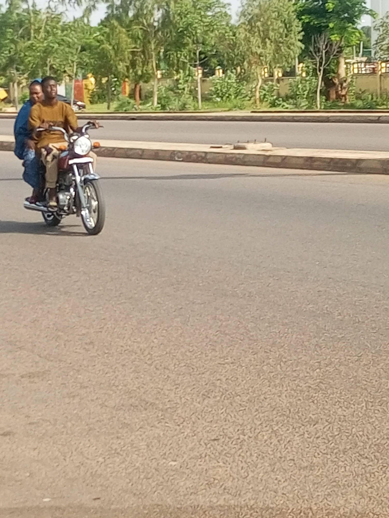 Two individuals are riding a motorcycle on a paved road in Gombe, Nigeria. The driver, a man wearing a brown shirt and tan pants, is in the front. A passenger, wearing a blue head covering and blue clothing, is seated behind him. The motorcycle is dark red with silver accents and chrome components. The road surface is asphalt. In the background, there is a sidewalk with low stone walls and several trees. The trees are deciduous, with light green foliage, and some appear to be ornamental. Behind the trees, a building with a textured wall and decorative light fixtures can be seen. The sky is overcast.