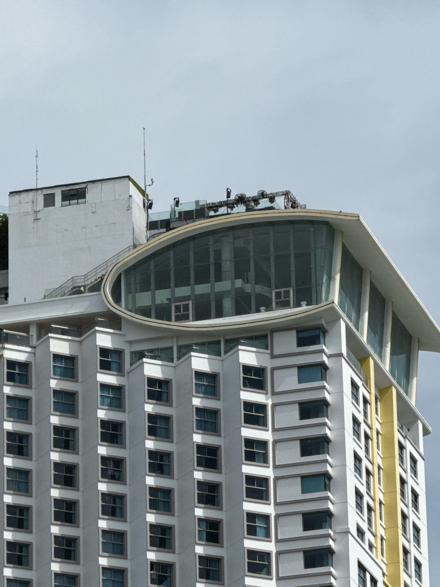This is a medium shot of a modern hotel building in Nha Trang, Vietnam. The building is predominantly white with a distinctive curved glass facade on its upper levels. The facade features an oval-shaped window that wraps around the corner of the building, offering panoramic views. The lower sections of the hotel are adorned with rows of windows, creating a repetitive geometric pattern. A bright yellow column is visible on the right side of the building, adding a splash of color to the otherwise neutral facade. The sky is overcast, suggesting a cloudy day. The top of the hotel features some external equipment, possibly for lighting or sound systems, hinting at an event space or rooftop venue. The overall impression is one of contemporary architecture designed to maximize natural light and views in a coastal city.