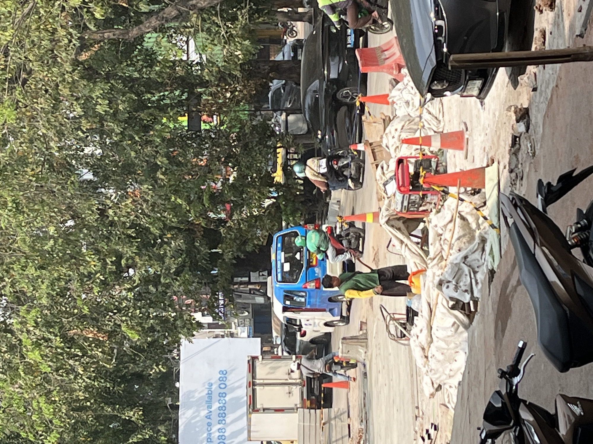 In the city of Unknown, Unknown, on a sunny day, a street scene unfolds with ongoing construction and traffic. Several motorcycles are visible, with one rider in a green helmet wearing a green jacket, and another rider wearing a tan shirt and a beige helmet. A man in a yellow and green shirt with black shorts stands near the construction area, holding what appears to be a broom. Construction materials, including white tarps and orange traffic cones, are scattered across the ground. In the background, various vehicles, including a white truck, a blue and white van, a black sedan, and other cars and motorcycles, navigate the street. Large trees with dense green foliage line the street, casting shadows on the ground. A large white banner with blue text advertising "Space Available" and a phone number is mounted on a building. The environment appears to be a typical urban street with a mix of residential and commercial elements.