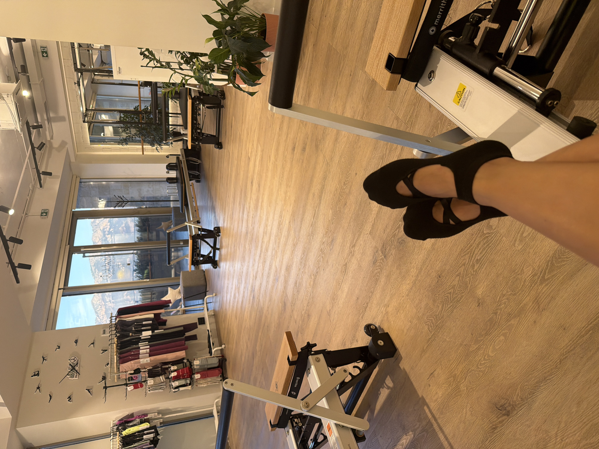 The image depicts the interior of a brightly lit Pilates studio, likely situated in Boreti, Montenegro. Sunlight streams through a large window, illuminating the polished wooden floor. The studio is equipped with multiple Pilates reformers, some in the foreground and others in the background. Clothing racks filled with workout attire and a display of socks are visible against a wall adorned with decorative clock and yoga pose figures. Potted plants add a touch of greenery to the space. In the foreground, a person's legs, wearing black Pilates socks, are casually resting on one of the reformers, suggesting a moment of relaxation after or before a session. The view through the window reveals a scenic mountain landscape, characteristic of the Boreti region.