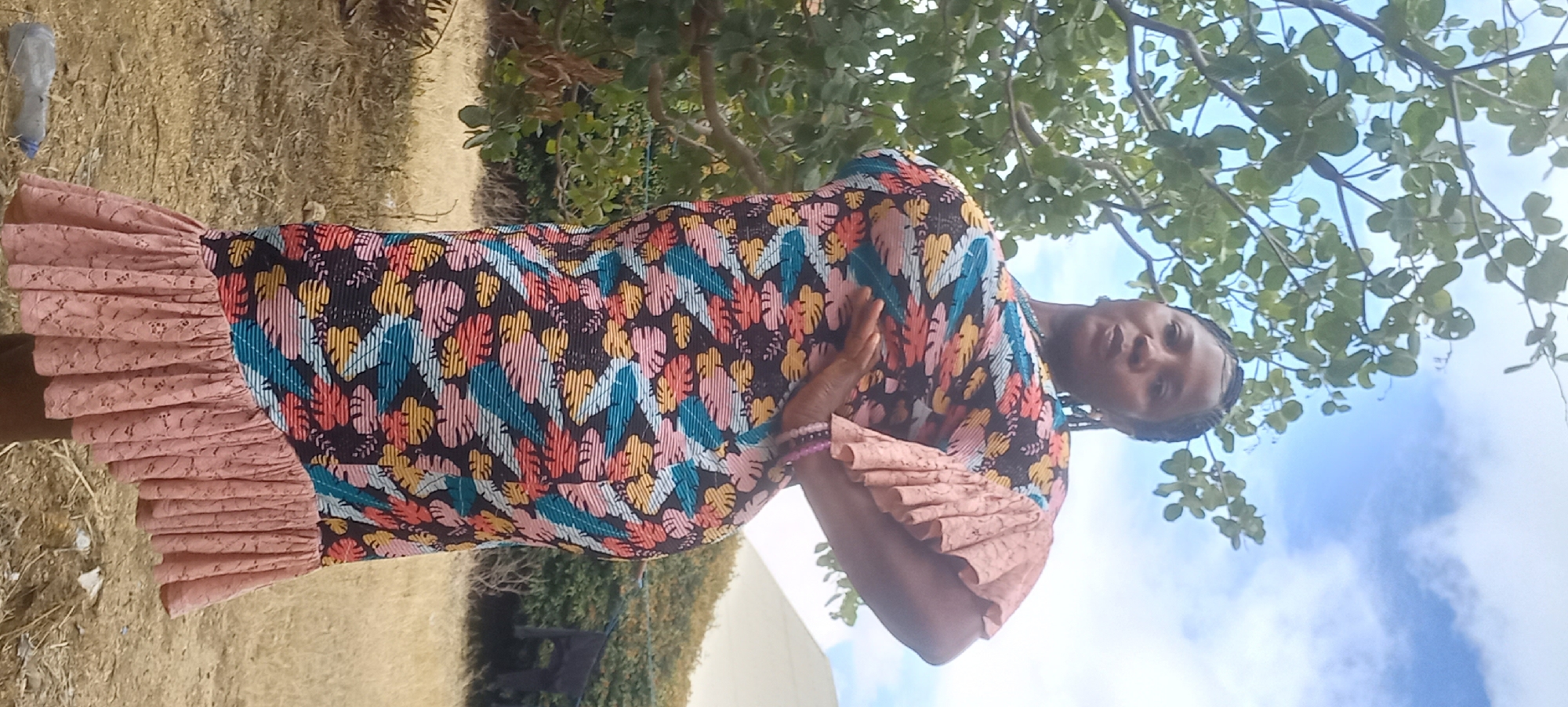 A Black woman poses for a photo outdoors with a tree and cloudy sky in the background. She is wearing a long, colorful, floral print dress with ruffled sleeves and a ruffled hem. Her hair is braided, and she has a bracelet on her left wrist. Her right hand is on her hip, and her left hand is resting on her stomach. The ground is covered in dry grass and some scattered debris. The overall scene appears to be in an outdoor, possibly rural, setting. The location in Jos, Nigeria, is not discernible from the image itself.