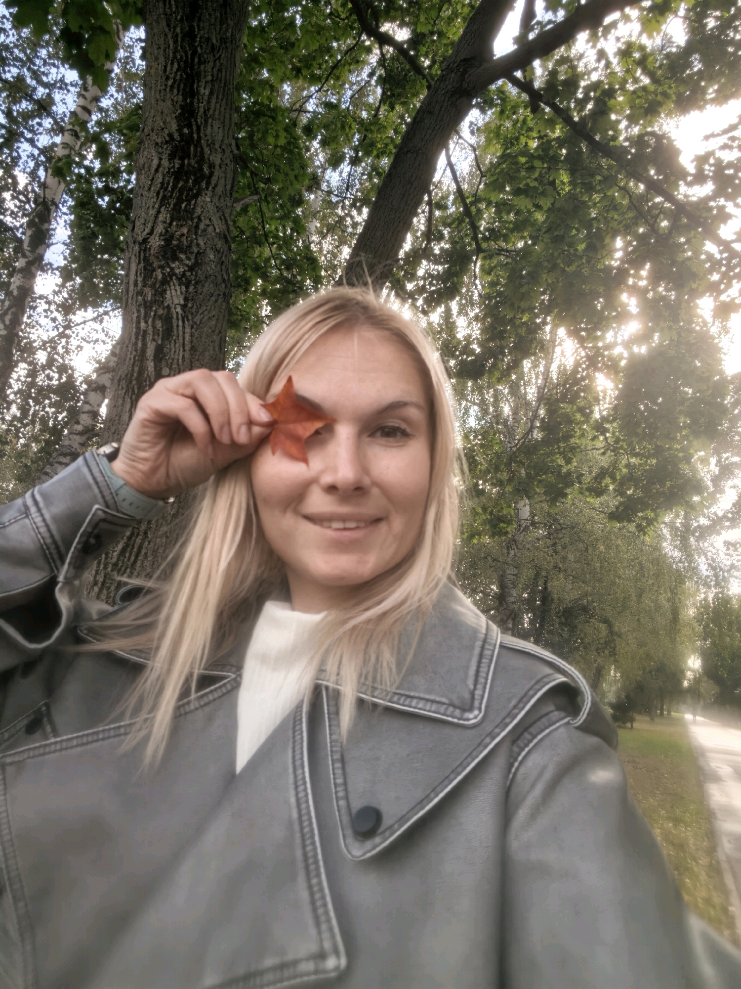 A woman with blonde hair is holding a red leaf in front of her right eye, obscuring it. She is smiling and looking at the camera. She is wearing a grey jacket with a white turtleneck underneath. The background shows trees and a path, suggesting an outdoor location. The scene appears to be in the city of Sterlitamak, Russia.
