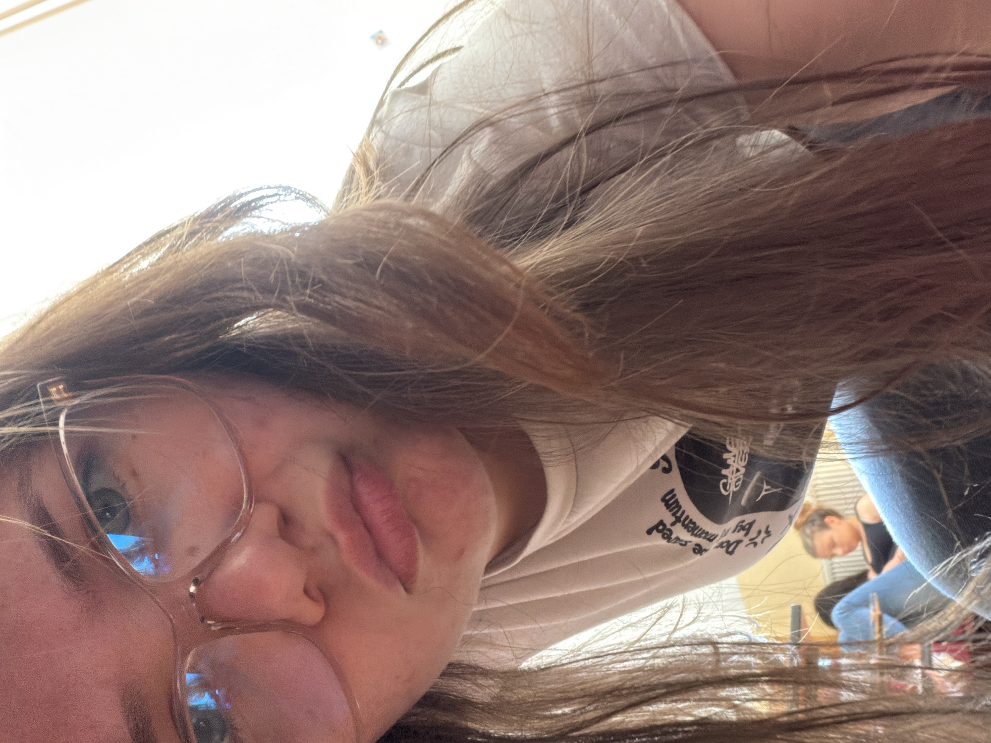 A selfie-style photograph depicts a young woman with long brown hair and eyeglasses. She is wearing a light-colored t-shirt with a black graphic design and text. The focus is on her face, with one eye and her lips clearly visible. The image is taken at an unusual angle, appearing tilted.
In the background, visible through gaps in the hair and clothing, is a glimpse of an interior setting. It shows another person, possibly female, wearing jeans and a dark top, seated or bending over. The interior appears to be a gym.
Given the lack of identifiable landmarks in the image and the reliance on subtle clues, determining a specific location in Veliko Tarnovo, Bulgaria is not possible. It can be said that the photograph was taken indoors, likely within a fitness center or gym facility in Veliko Tarnovo.