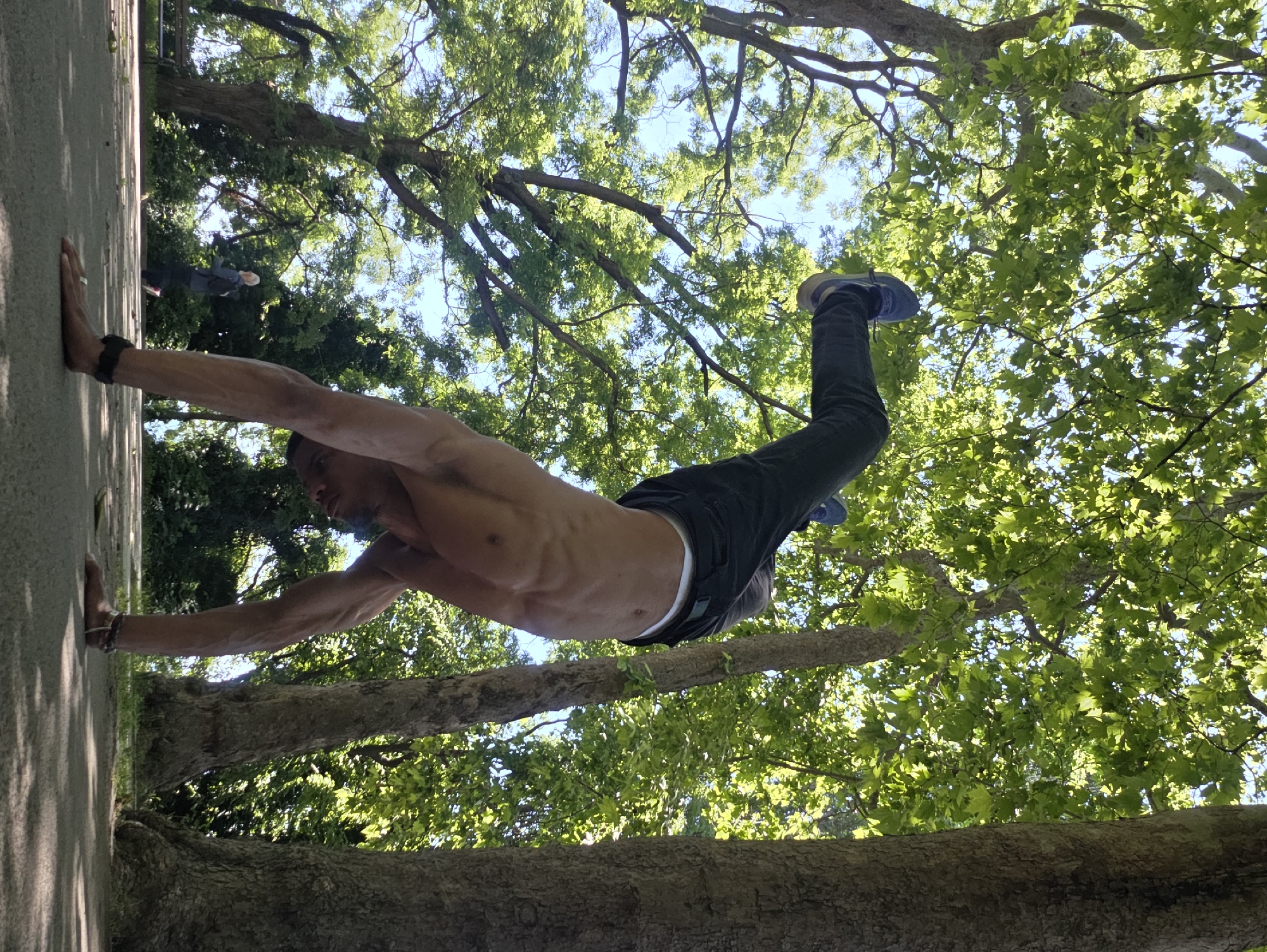 In a park in Varna, Bulgaria, a shirtless man is performing a handstand on a paved path. His legs are extended upwards, and he is wearing dark pants and blue and grey sneakers. The sun is shining through the leaves of large trees, creating dappled shade on the ground. In the background, another person is walking, and a park bench is visible. The man's abdominal muscles are prominently displayed.
