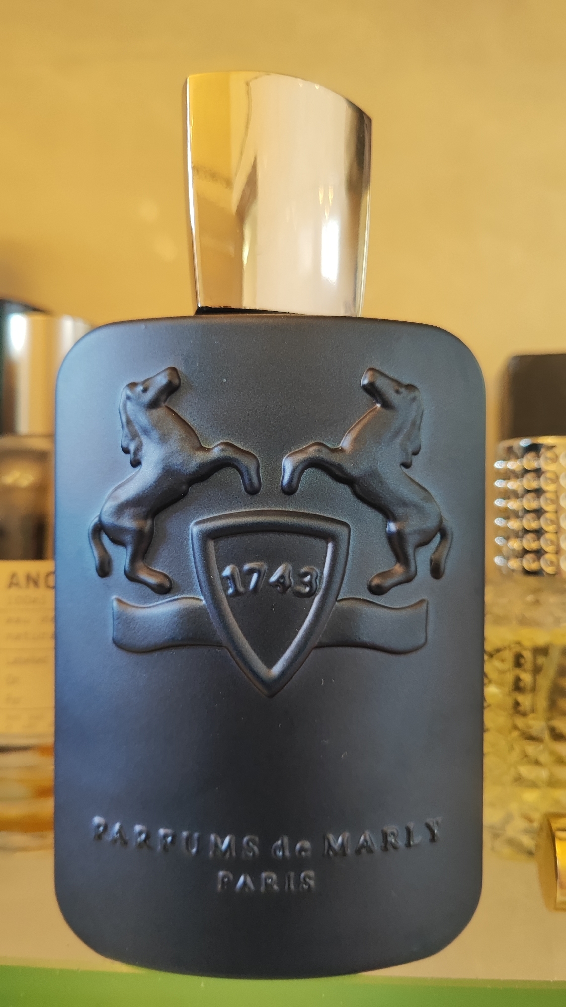 A black perfume bottle with embossed designs is centered in the frame, against a blurred yellow background. The bottle features two embossed horses facing inward, a shield with the number "1743" inside, and text at the bottom that reads "PARFUMS de MARLY PARIS." The cap of the bottle is silver and reflective. The scene is an indoor setting, possibly a shelf or display, with other blurred objects visible in the background. There is no indication of the city of Sochi, Russia, or any people.