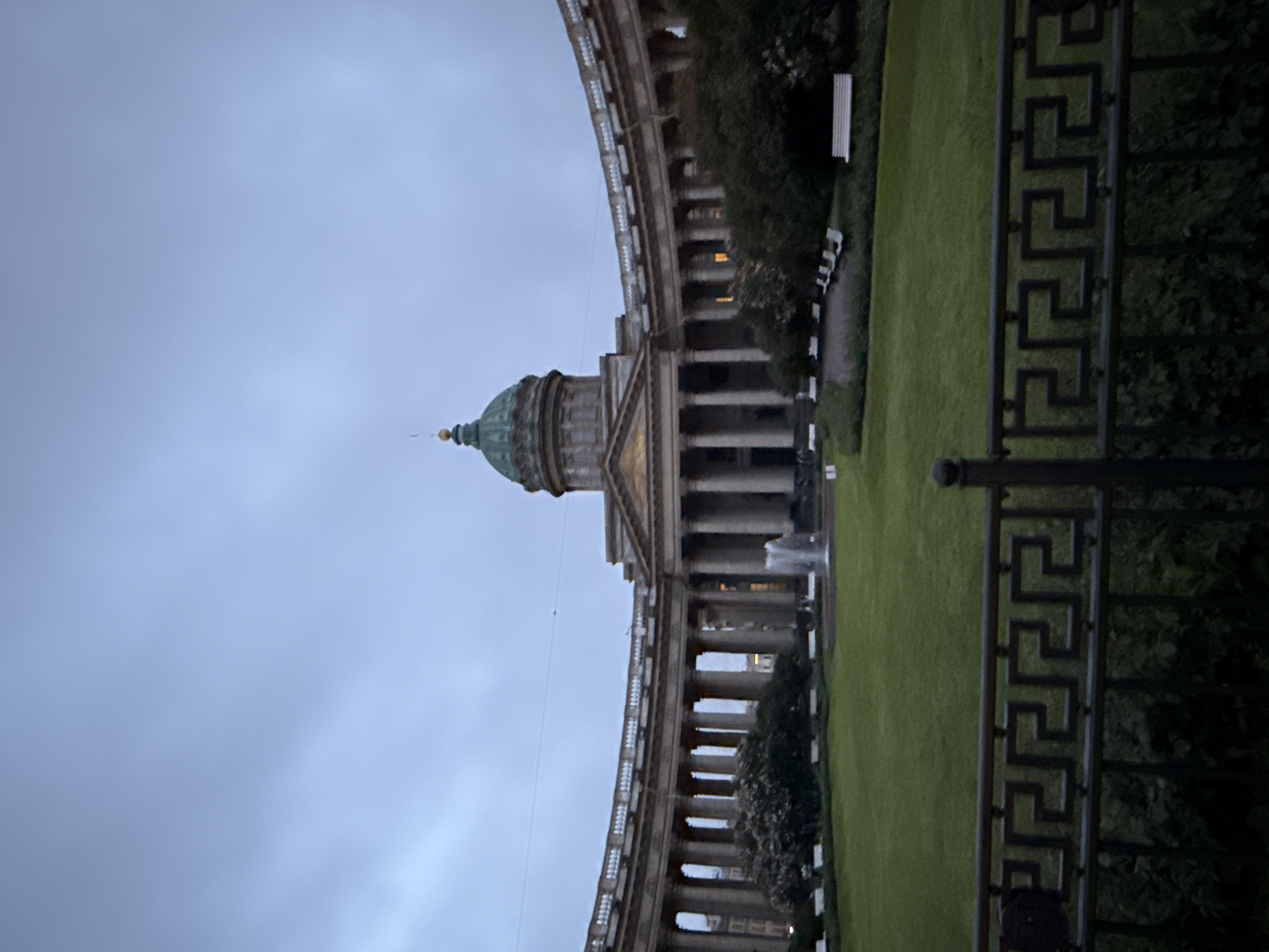 The image shows Kazan Cathedral, a prominent landmark located on Nevsky Prospekt in Saint Petersburg, Russia. The cathedral features a distinctive semicircular colonnade with numerous classical columns, enclosing a central courtyard. In the center of the courtyard, a fountain sprays water upwards. Beyond the colonnade, the main body of the cathedral is visible, topped by a large green dome. The sky is overcast and grey, suggesting it is either early morning, late evening, or a cloudy day. The foreground of the image is partially obscured by a dark metal fence with an intricate Greek key pattern. Several white benches are visible on the green lawn within the courtyard. Trees and bushes are strategically placed around the perimeter of the courtyard. The overall scene depicts a quiet and serene atmosphere, with no people clearly visible.
