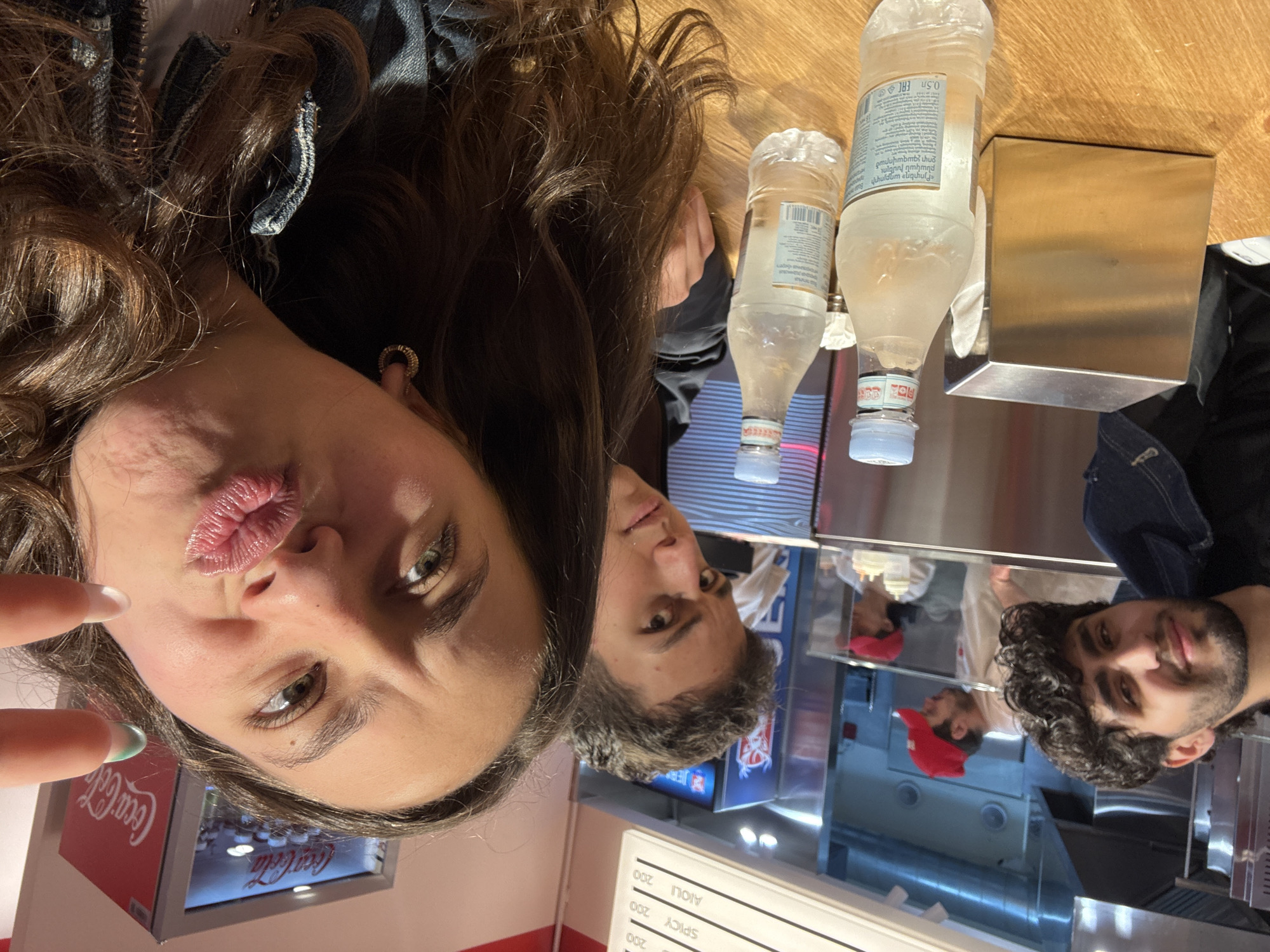 This portrait-oriented image, taken from a high angle and capturing a selfie, depicts three individuals in what appears to be a casual eatery or food stall. The primary subject is a woman in the immediate foreground, with long brown hair cascading down, making a playful puckered-lip "duck face" expression towards the camera. She has visible earrings and light-colored nail polish on her fingers, one of which holds the phone.
Behind her, another person with dark hair is partially visible, looking slightly away from the camera. In the mid-ground, seated or standing behind a wooden counter, is a man with dark, curly hair and a beard, looking towards the camera with a subtle smile.
On the wooden counter in front of the subjects are two plastic water bottles. Their labels feature Armenian text, including "«Բյուրեղ» աղբյուրի բնական խմելու ջուր չգազավորած" (Byuregh spring natural drinking water, non-carbonated) and "0.5л ERC", while the blue cap of one bottle clearly shows "Evian". Next to the bottles is a reflective, cuboid metallic dispenser.
The background reveals a service area or kitchen with shiny, metallic surfaces, suggesting a commercial food establishment. Reflections of people, including one person wearing a red cap, are visible in these surfaces. To the right, a white menu board lists items such as "AIOLI 200" and "SPICY 200," with "200" likely indicating a price. A "Coca-Cola" sign is also visible on a display in the lower left. The overall lighting suggests an indoor setting, possibly during the day or evening. The visible Armenian text on the water bottles and the provided context confirm the location as Yerevan, Armenia.