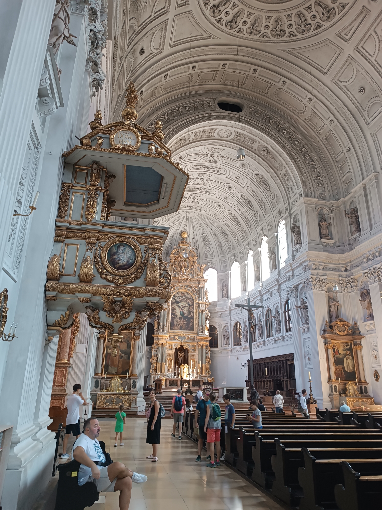 This is an interior view of the Frauenkirche, also known as the Cathedral of Our Dear Lady, in Munich, Germany. The image captures the grandeur of the cathedral's nave, with its high, vaulted ceilings adorned with intricate stucco work. Sunlight streams through arched windows, illuminating the ornate pulpit on the left and the main altar at the far end of the church. Several people are present, observing the architecture and engaging in various activities. In the foreground, a man is seated on a chair, looking to the left. Further into the nave, other visitors are seen walking and standing, some in small groups. The pews of the church are visible, with a few individuals seated and others standing near the altar. The overall impression is one of a serene and majestic religious space, with visitors exploring its historical and architectural significance.