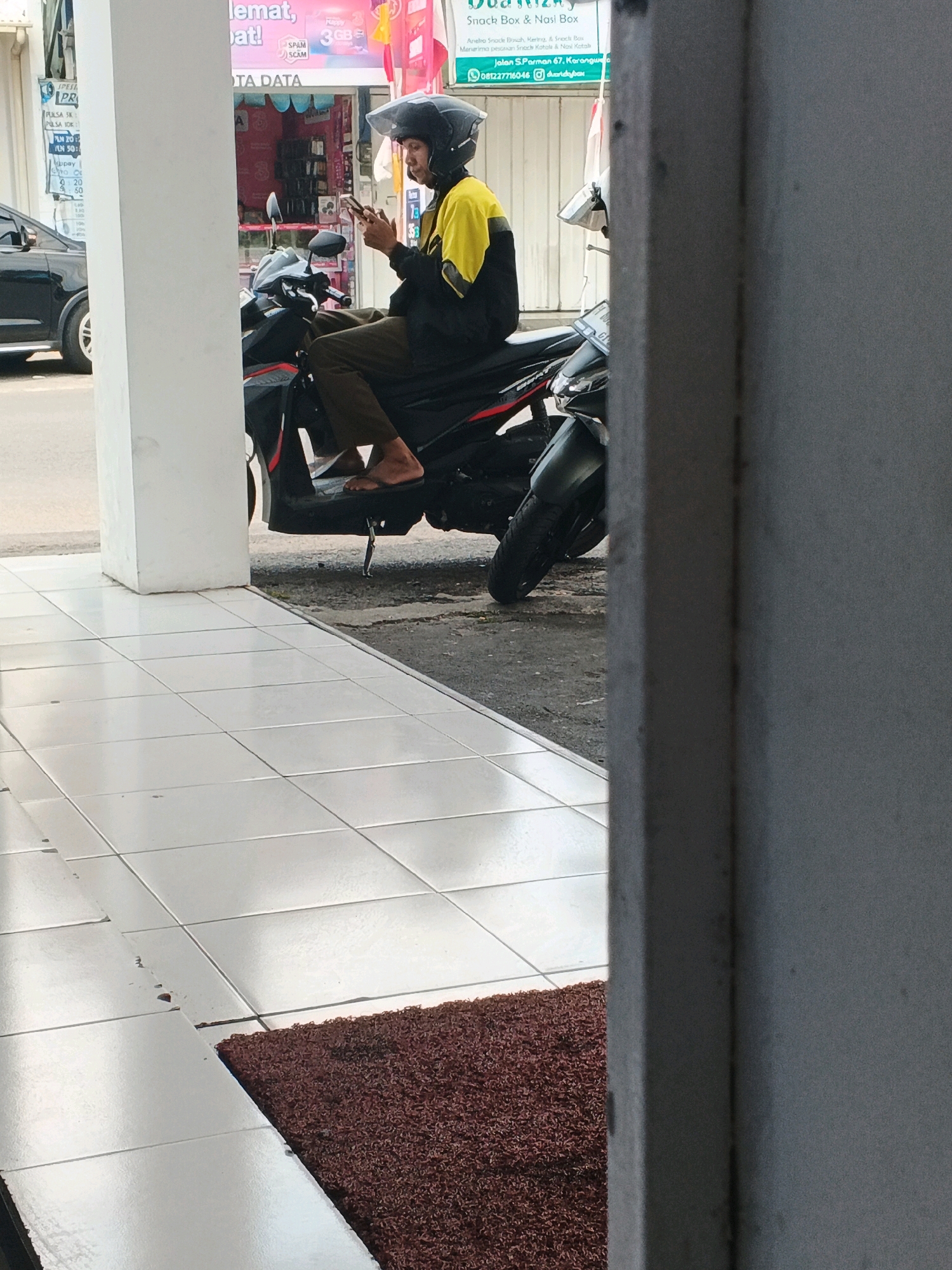 A person wearing a black and yellow jacket and a helmet is sitting on a motorcycle and looking at their phone. The motorcycle is parked on a street next to a building with shops. The person is wearing dark pants and flip-flops. The background shows the facade of a shop with various signs and advertisements, including one that appears to be for "DATA DATA" and another that says "SPAM SCAM." A black car is visible parked on the street to the left of the motorcycle. The ground in front of the building is tiled. The image appears to be taken during the daytime.