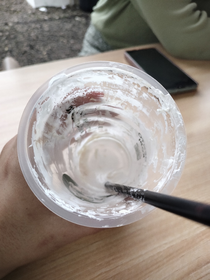 A close-up, top-down perspective captures an almost empty clear plastic cup, held by a person's hand visible on the left. The inside of the cup is coated with white, creamy residue, suggesting a beverage with a cream or whipped topping has been mostly consumed. A dark-colored straw or long spoon extends into the cup, reaching the bottom where a small amount of liquid or residue remains.
The background is blurred, revealing a light wooden table surface across the lower and right sides of the frame. On the table, a dark smartphone with a dark case lies horizontally. Behind the phone, partially visible, is another person wearing light green or olive-colored clothing. The far left background shows a highly out-of-focus outdoor area with textures resembling gravel or uneven ground.
The scene suggests an outdoor or semi-outdoor dining or cafe setting in Dramaga, Indonesia, during the daytime, given the natural lighting. No specific emotions are evident, and any text on the cup is too faint and blurry to be legible.