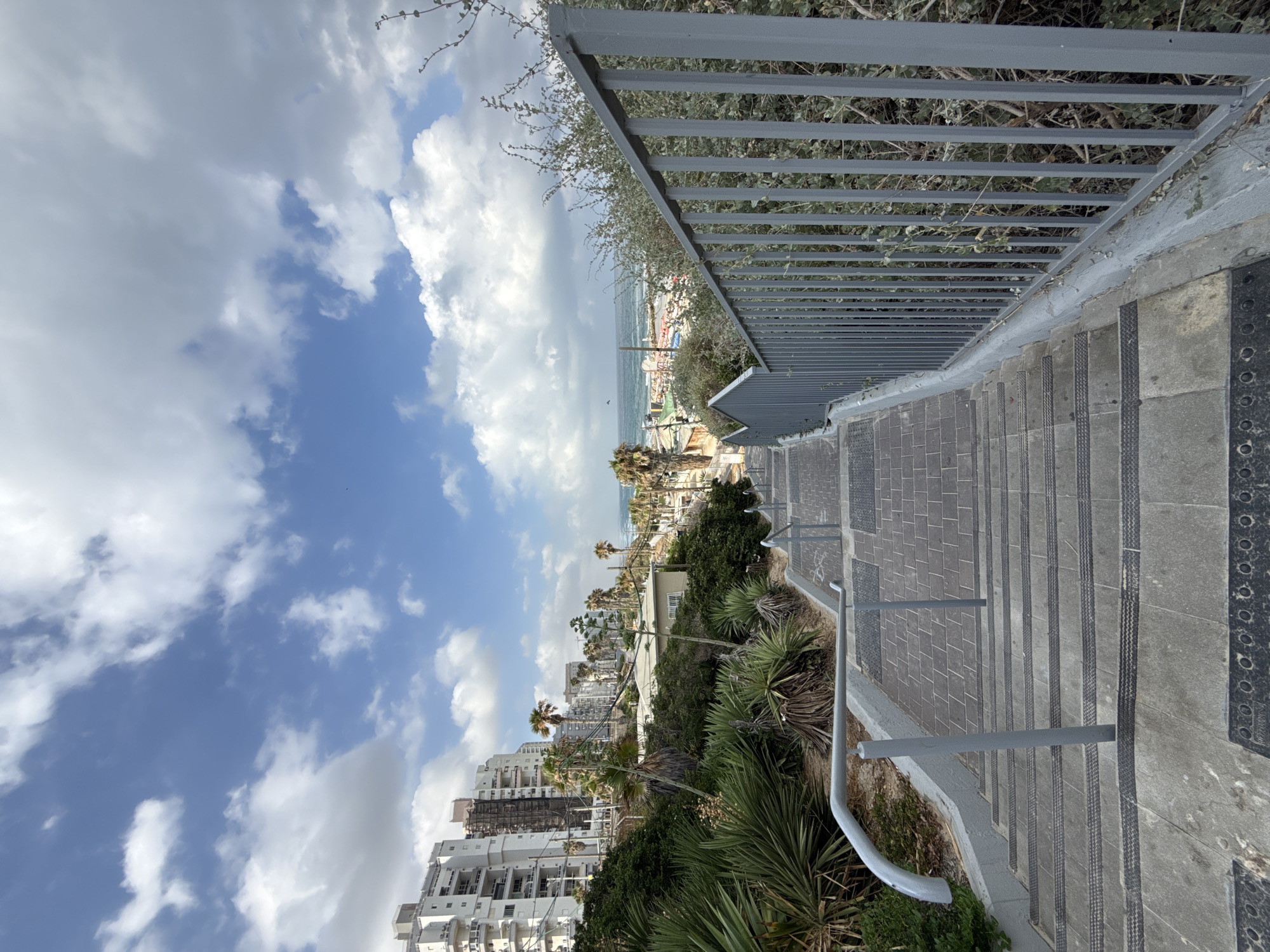 A downward view of a wide, paved staircase with gray concrete steps and darker textured treads. Metal handrails, painted gray, run along both sides of the staircase and also down the center. To the left of the stairs is a sloped area with lush green vegetation, including large agave plants and smaller bushes. To the right, a gray metal fence with vertical bars separates the stairs from more dense green shrubbery. In the background, several modern, multi-story white apartment buildings are visible on a hillside. Palm trees are scattered among the buildings and along a road that leads down towards a body of water, likely the Mediterranean Sea, which is visible in the distance. A small beach with scattered beach chairs and umbrellas can be seen near the water's edge. The sky is a vibrant blue with scattered white cumulus clouds. The overall scene suggests a coastal urban environment.