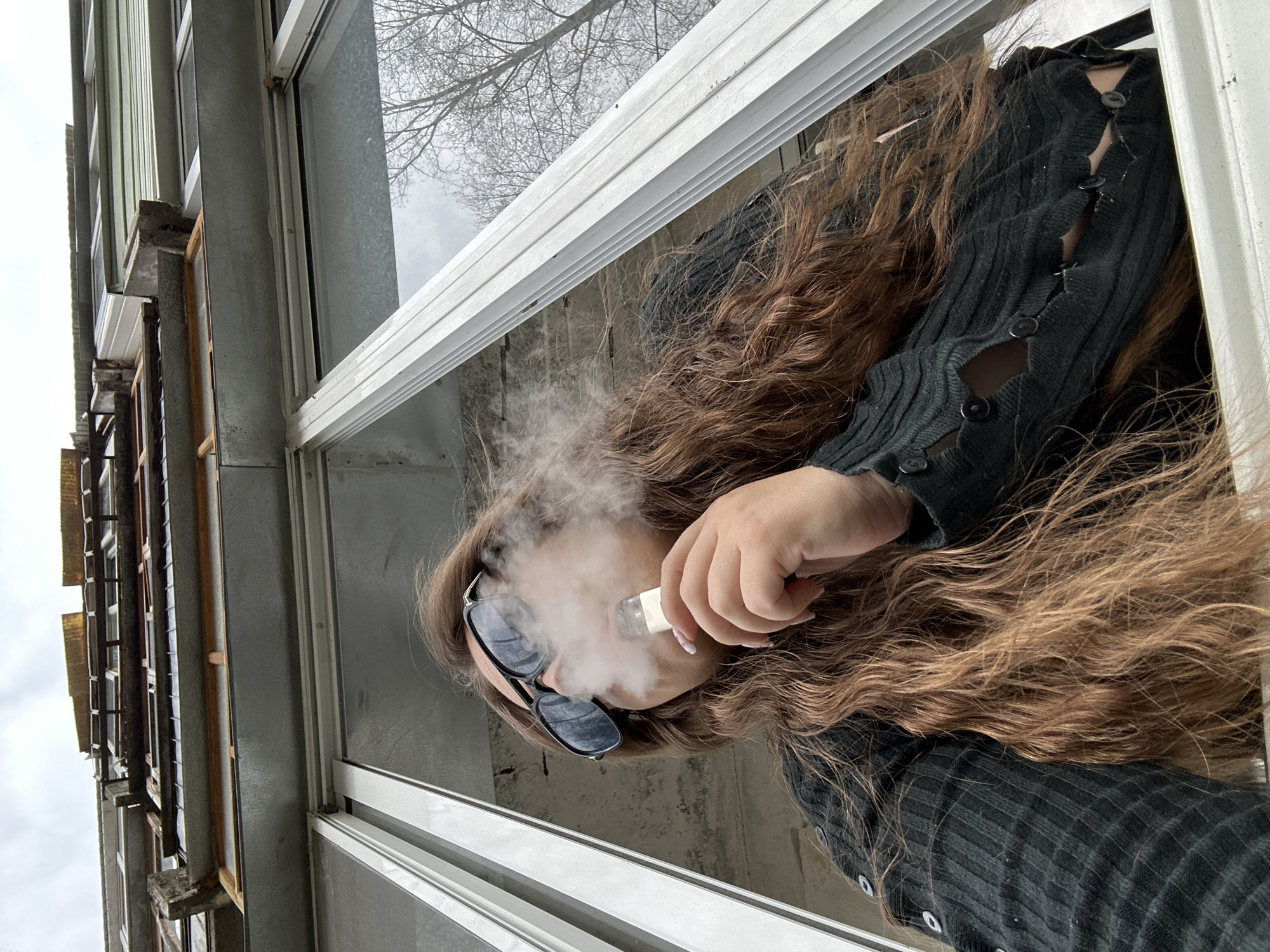 A woman with long, wavy brown hair is pictured from a low angle, presumably looking out a window. She is wearing sunglasses and a black, button-down sweater. She is holding an object to her mouth that is emitting smoke. The background shows a building exterior with multiple windows, suggesting an urban setting. Tree branches are visible through the window, indicating it is possibly outdoors. It is difficult to ascertain the specific location in Chaikovsky, Russia, without more specific details.