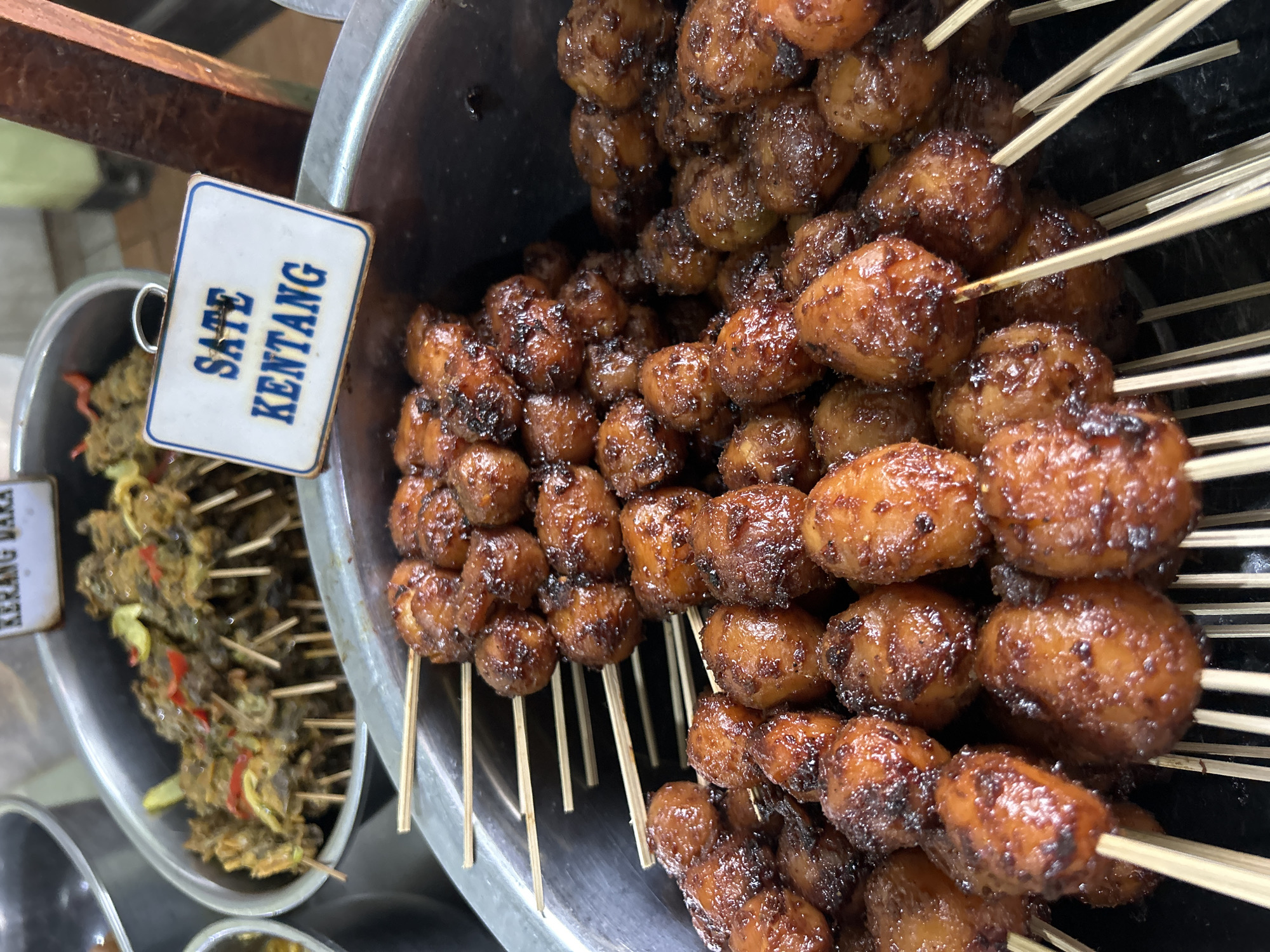 The image shows a close-up view of several skewers of "Sate Kentang" (potato satay) in a large metal container. The potatoes are small, round, and appear to be glazed with a dark, sticky sauce, some pieces showing charring from grilling. They are arranged on wooden skewers. A white sign with blue text reading "SATE KENTANG" is visible in the upper right portion of the image. Behind the potato satay, a similar metal container holds skewers of what appears to be "Kerang Dara" (blood clam) satay, identifiable by the darker, less uniform shapes and specks of red and yellow ingredients. The scene is likely a food stall or market in Cirebon, Indonesia, a city known for its diverse street food offerings, particularly satay variations. The lighting suggests an indoor or covered outdoor setting, possibly a market during daylight hours. There are no people visible in the frame.