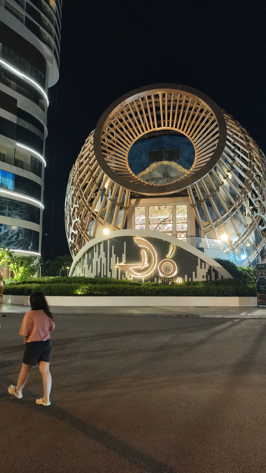 An outdoor night-time photograph captures a woman walking in front of a striking, spherical building with a latticed wooden exterior. The building, illuminated by a warm glow, features a prominent illuminated sign that reads "DO Theatre". To the left of the spherical building stands a tall, modern cylindrical building with horizontal bands of lights. The foreground is a dark asphalt road with faint tire tracks and shadows. The background is a dark night sky. The woman is seen from the back, wearing a pinkish-orange t-shirt and dark shorts, with her legs slightly apart as she walks. She has long, dark hair. The overall setting is urban.