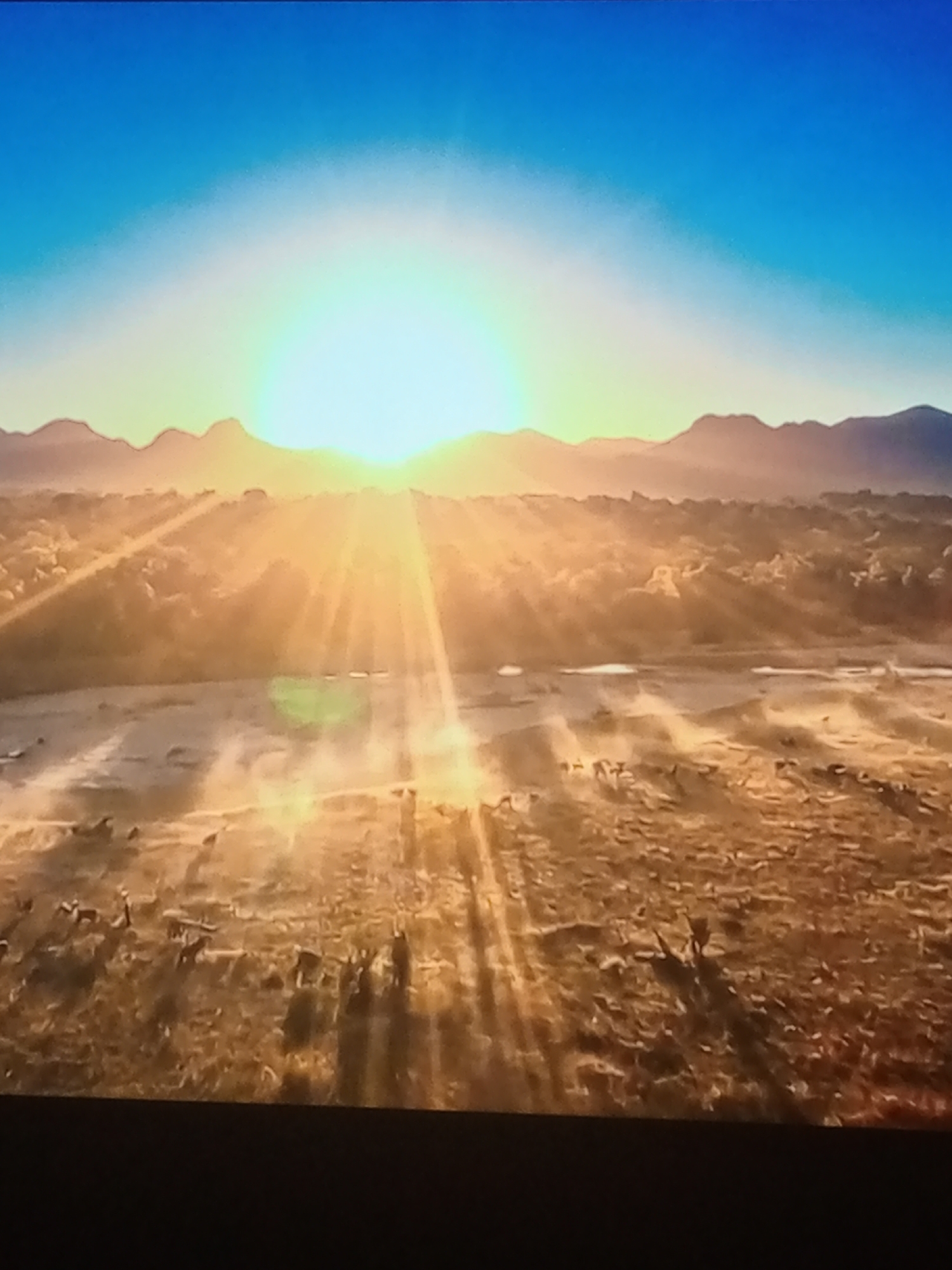 An aerial, medium shot captures a vast savanna landscape bathed in the warm, golden light of a setting sun. The sun, positioned near the center of the frame, casts long, dramatic rays across the scene, illuminating dust kicked up by a herd of animals moving across the dry, grassy terrain. In the distance, a range of mountains forms a silhouetted horizon against a gradient sky that transitions from a vibrant blue at the top to a soft yellow near the sun. Numerous animals, appearing to be wildebeest or similar ungulates, are scattered throughout the middle ground and foreground, some kicking up dust as they traverse the landscape. The shadows of these animals stretch long, emphasizing the low angle of the sun. The overall impression is one of a natural, untamed environment at dusk.