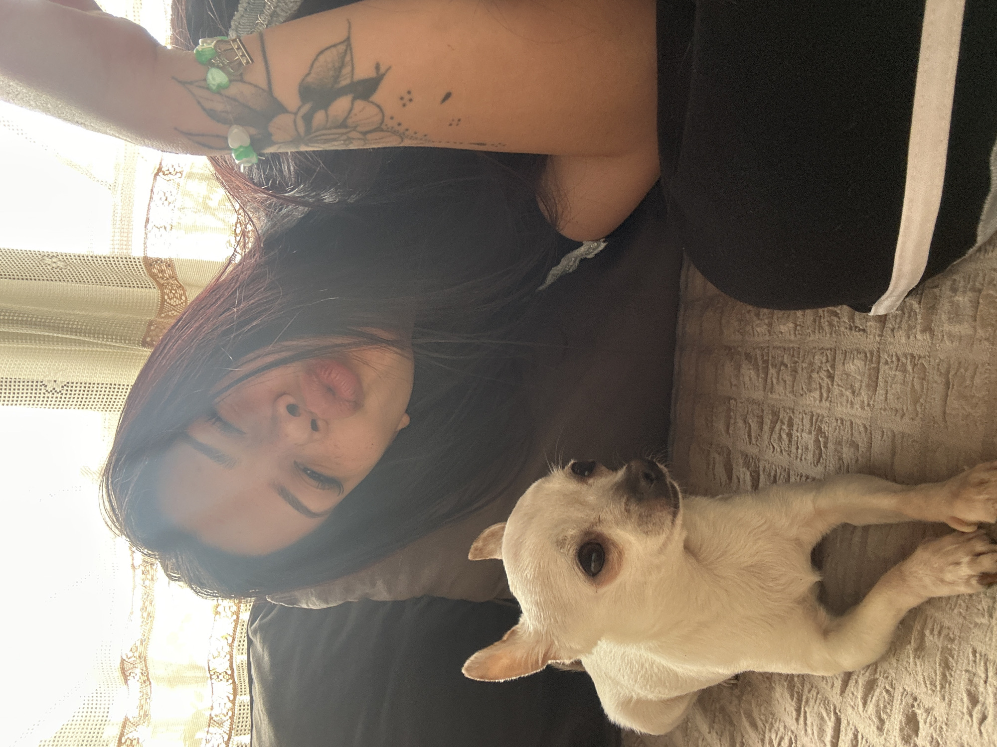 A woman with dark hair lies on a brown textured sofa next to a small, white dog with short hair. The woman wears black pants with a white stripe down the side and a bracelet on her right wrist, which has a tattoo of a flower and three dots. She is facing the camera. The dog is lying on its stomach, looking up at the camera. A lace-edged curtain is visible in the background. The location is unconfirmed.