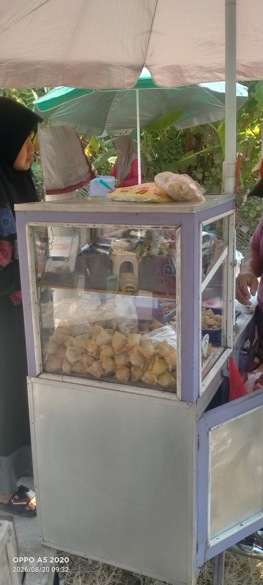 The image depicts a street food stall, likely in Sowan Kidul, Indonesia, with two women visible. The stall features a glass display case filled with numerous small, fried pastries. Above the glass, there are two sealed plastic bags of food items. One woman, wearing a black hijab, is on the left side of the frame, facing the stall. Another woman, wearing a light brown hijab and a red top, is visible in the background behind the stall, appearing to be engaged in some activity related to the stall. The environment is outdoors, under what appears to be a large umbrella providing shade, with foliage visible in the background, suggesting a tropical or subtropical setting. The overall scene is illuminated by natural daylight.