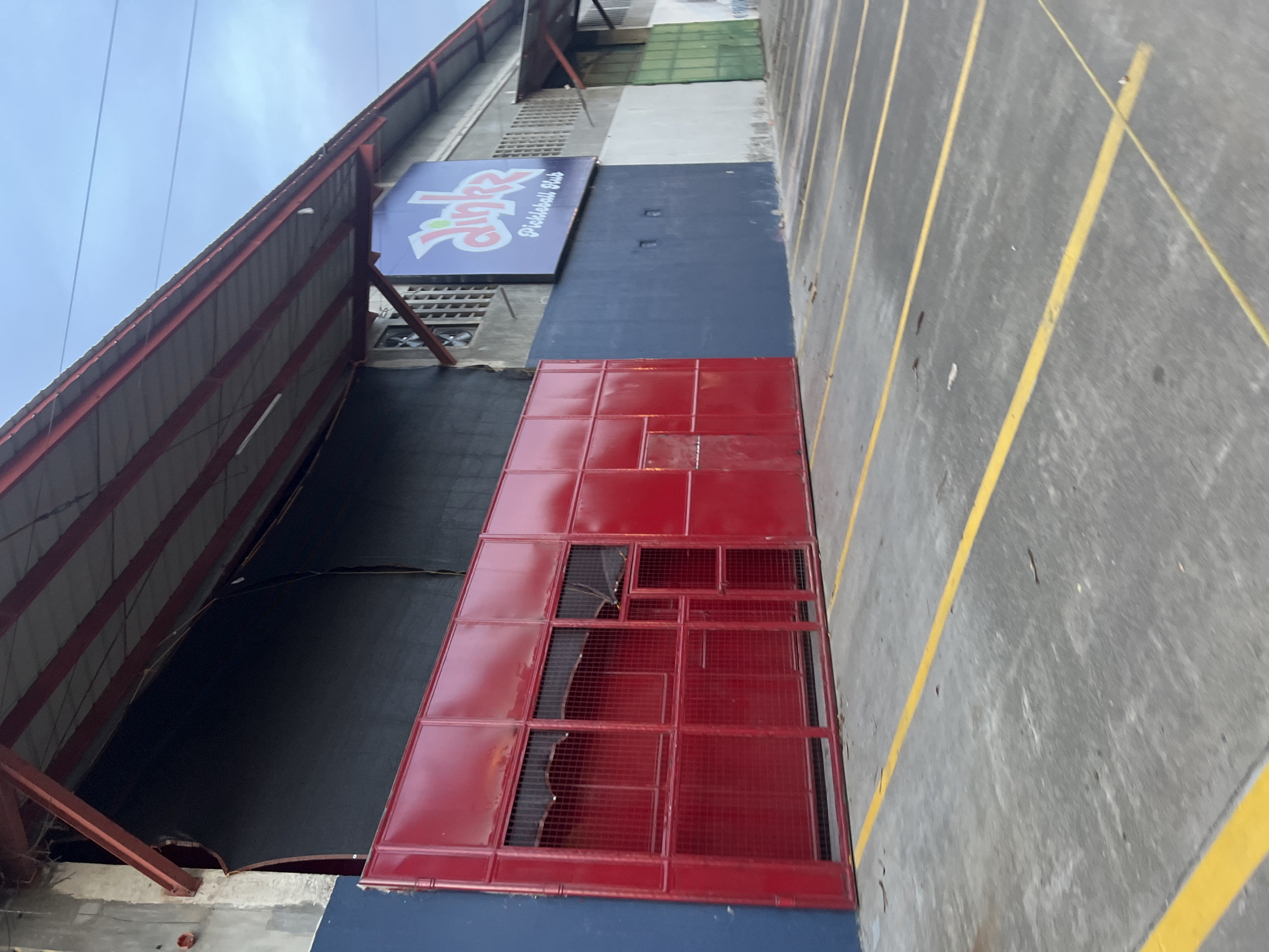 This is a medium-angle, eye-level outdoor shot of a commercial building. The building has a dark gray, corrugated metal roof supported by red steel beams. A prominent feature is a large, red metal gate with wire mesh infill, which appears to be partially damaged. The walls of the building are painted in sections: a dark blue section to the left of the gate and a lighter gray section to the right, beyond which are sections of white and green painted walls. Above the dark blue wall, a purple sign with pink lettering reads "dinkz Pickleball Club." The ground in front of the building is a concrete parking lot marked with yellow lines, indicating parking spaces. The sky above is overcast. This location is the "dinkz Pickleball Club" in Davao City, Philippines.