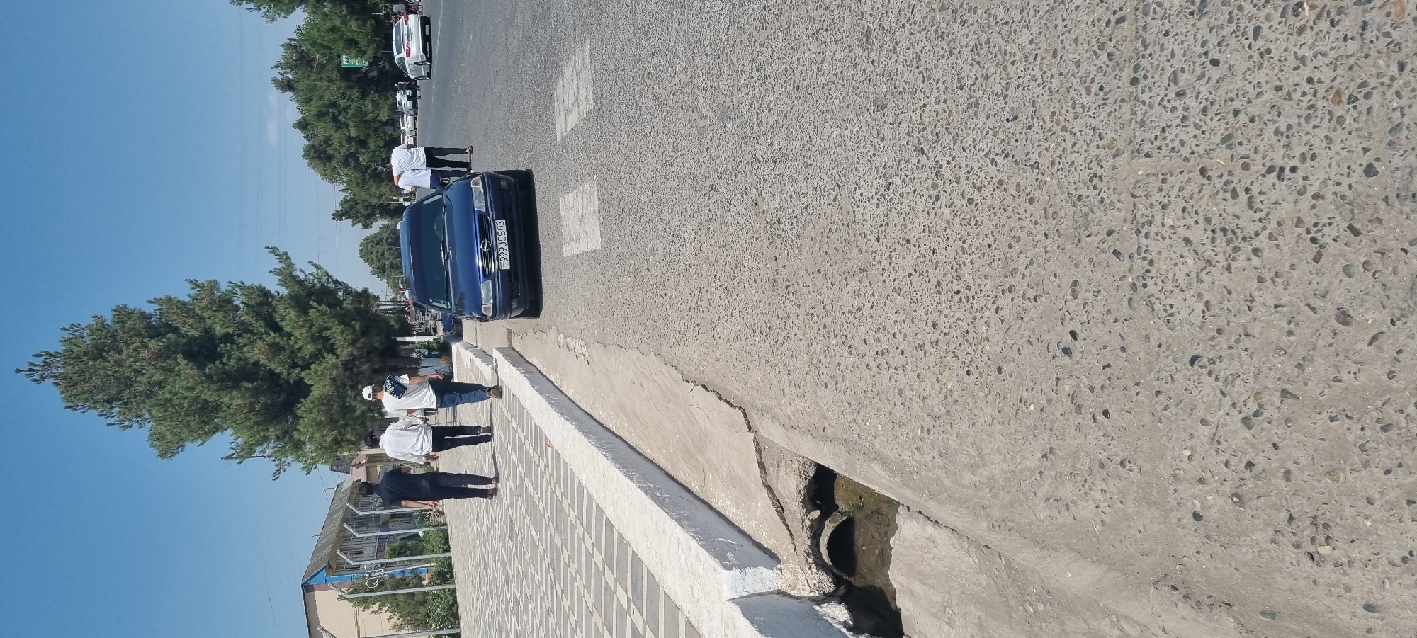 In Ismoili Somoni, Tajikistan, a blue Opel Astra with license plate 999OS503 is parked on the side of a street. The car is positioned at a crosswalk, with its front wheels slightly over the white lines. In the background, several people are walking along a paved sidewalk next to a tree-lined street. Buildings are visible on the left side of the street. A drainage ditch runs along the edge of the sidewalk, with a metal pipe visible inside. The sky is clear and blue.