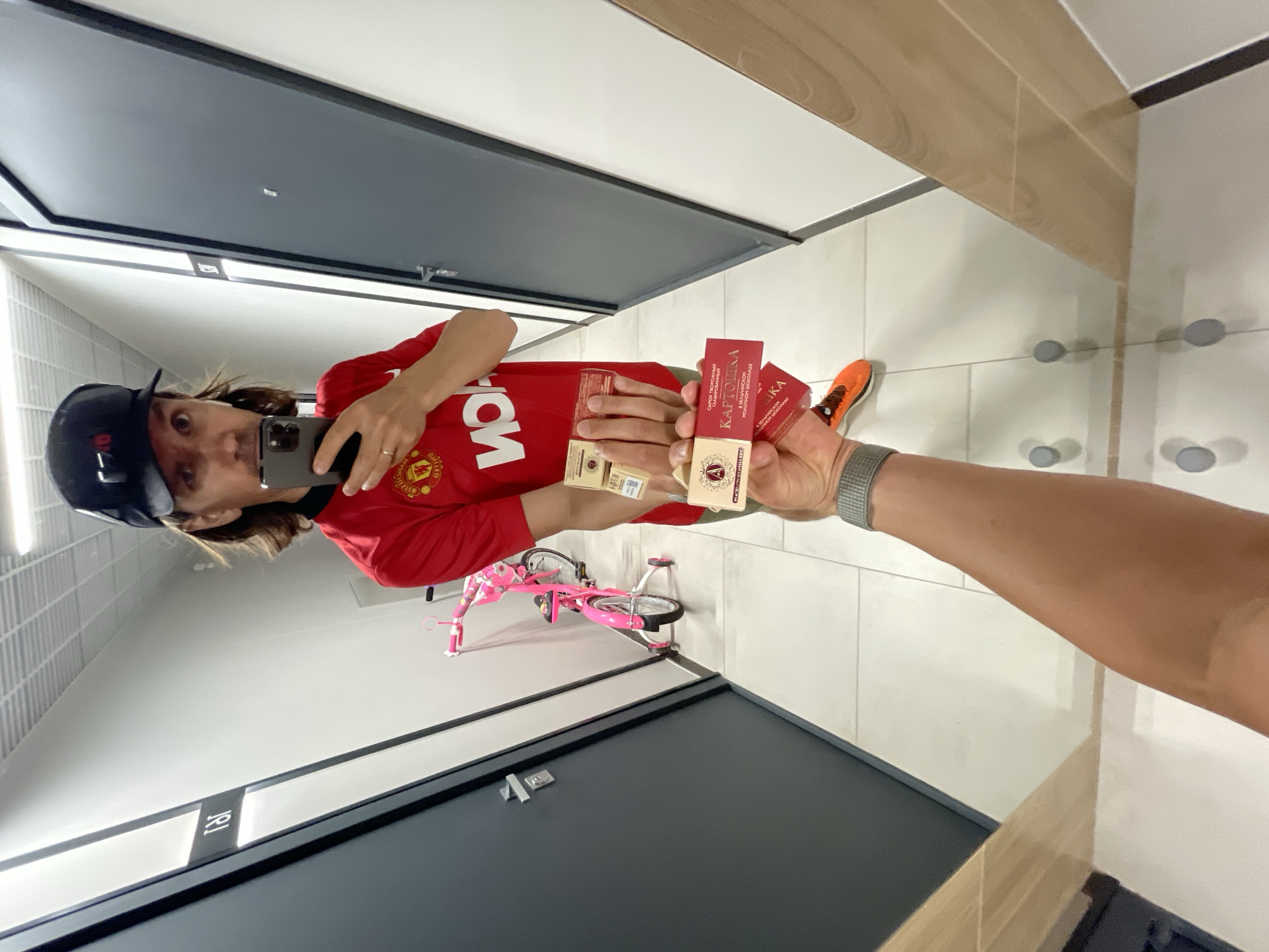 A person wearing a red Manchester United shirt and a black baseball cap is reflected in a mirror, taking a selfie with a smartphone. They are holding multiple packages of "Картошка" (Kartoshka), a Russian chocolate snack, in their hands. A pink child's bicycle is visible in the background, and the surrounding environment appears to be a tiled hallway or restroom with grey doors. An orange running shoe is partially visible as well. The specific location is Unknown, Unknown.