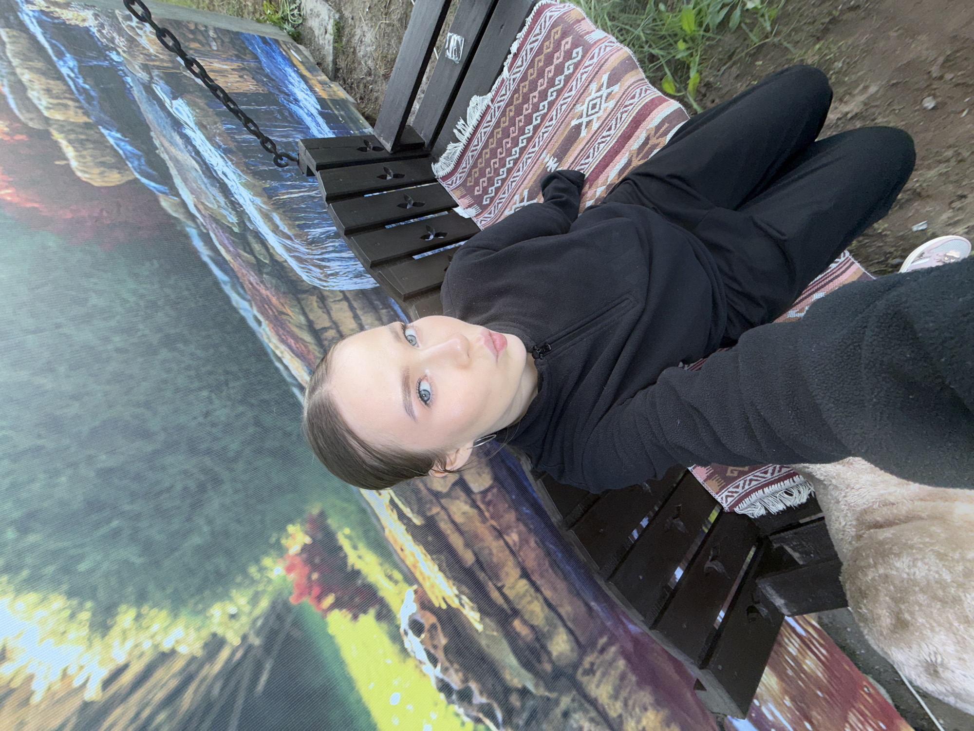 A young person with light brown hair pulled back is lying on a dark wooden bench or swing, looking directly at the camera. They have light blue eyes and a calm, neutral expression. The person is dressed in a black fleece jacket with a zipper and black pants.
They are resting on a patterned blanket or rug draped over the bench, featuring geometric designs in shades of red, white, brown, and pink. The bench itself is made of dark-stained wooden slats. A heavy black metal chain, possibly part of the swing mechanism, is visible in the upper left quadrant of the image.
To the left of the bench, a large, vibrant banner or tapestry is laid out, displaying a colorful, abstract, or natural landscape design with prominent blues, greens, yellows, and reds, possibly depicting waterfalls or stratified rock. The ground around the bench appears to be dirt and sparse green grass. The lighting suggests it is daytime. There is no visible text in the image.