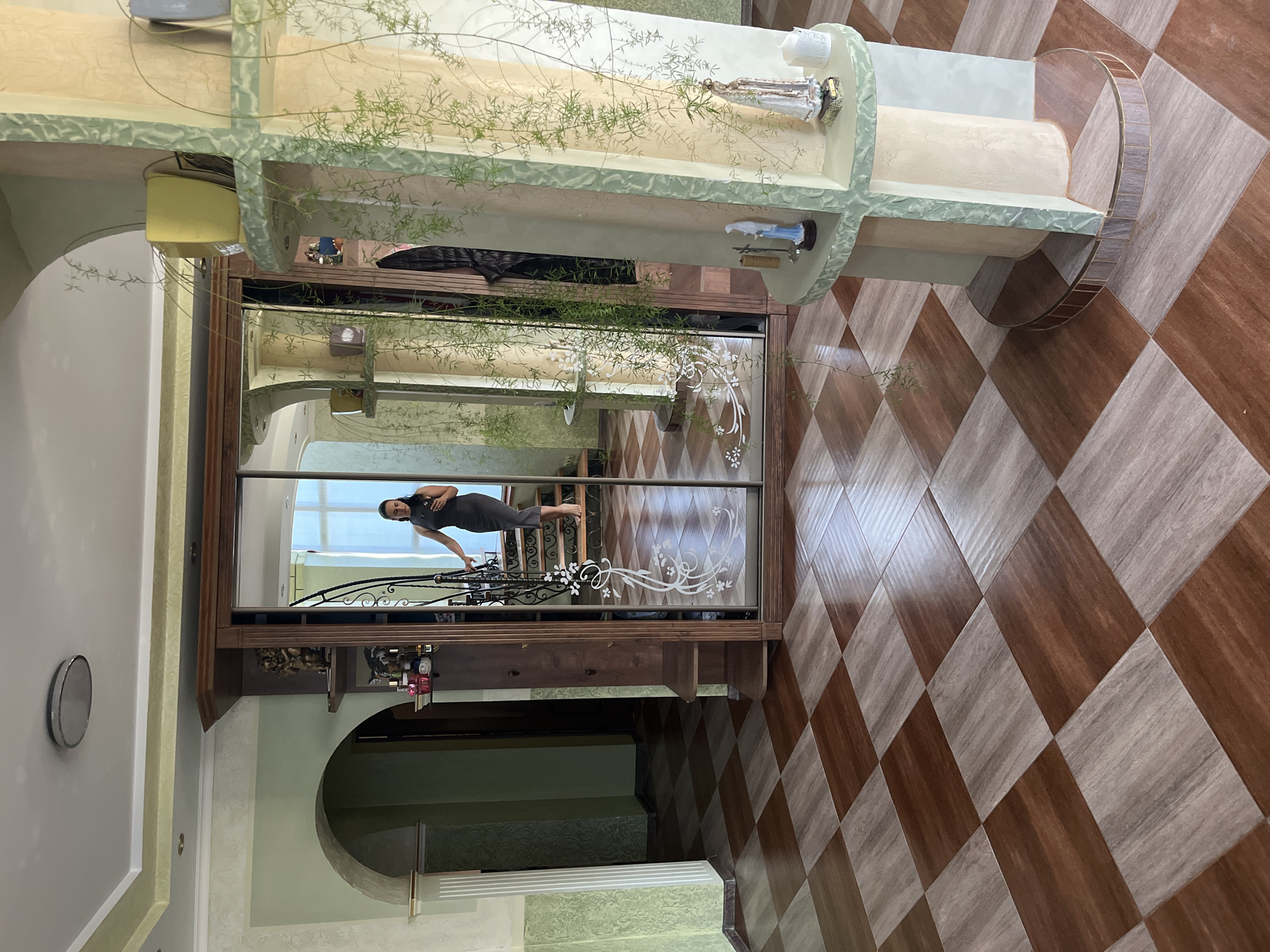 A full shot captures a residential interior with a checkered floor of dark brown and light beige tiles, leading towards a large wooden wardrobe with mirrored doors. A woman in a dark grey dress, her dark hair pulled back, is reflected in the mirror, standing on a staircase with black metal railings decorated with white floral designs. She is holding a smartphone in her left hand. To the left of the wardrobe, an arched entryway with light green walls leads into a darker hallway. To the right, a decorative pillar, also in light green and beige tones, features multiple shelves with potted plants, figurines of saints, and a candle. The walls are painted in light green and beige, with textured detailing. The ceiling is white with a central circular light fixture. The environment appears to be a well-lit hallway or entryway of a home.