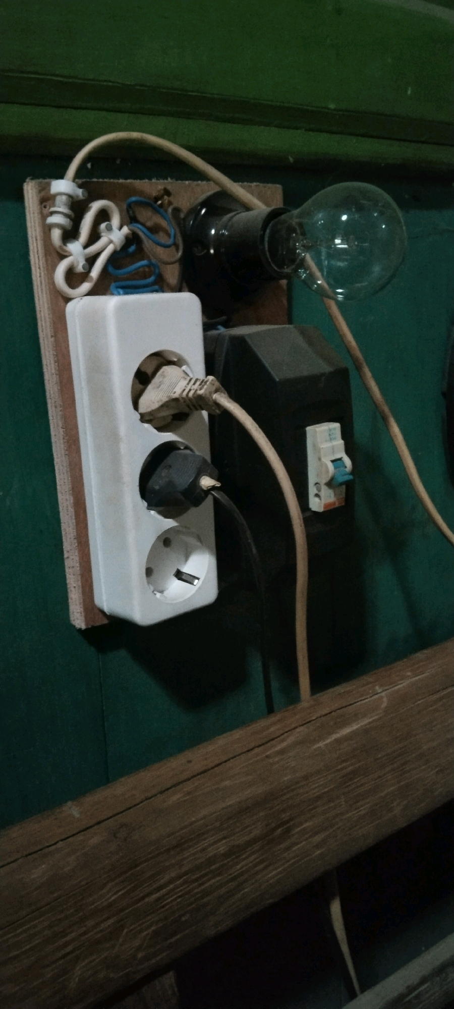 A close-up, vertical, eye-level shot shows an electrical outlet strip with a light bulb, a circuit breaker, and various electrical wires and plugs. The outlet strip is white and has two sockets, one with a plug inserted. Another plug is inserted into the second socket. The light bulb is clear and is screwed into a black socket. A circuit breaker, white with a blue toggle, is mounted in a black housing. The electrical components are mounted on a piece of wood, which is attached to a green wall. A wooden beam runs across the bottom of the image. The scene appears to be in a dimly lit, possibly unfinished, indoor space. The location is described as Wadang, Indonesia.
