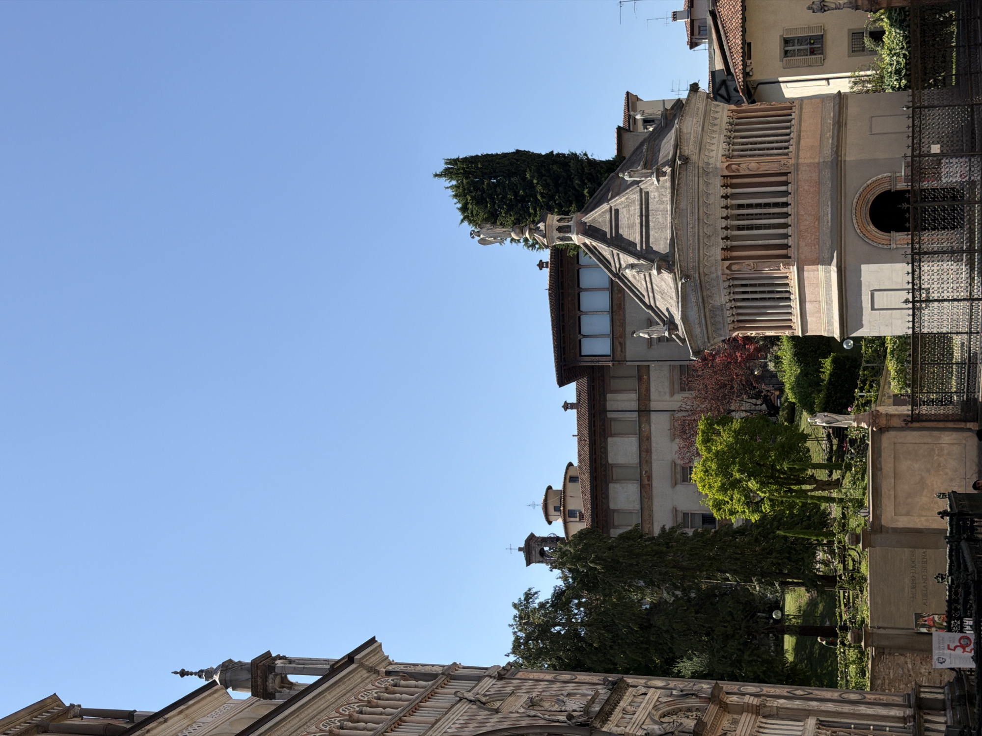 The image captures a daytime scene in Mozzo, Italy, likely in an urban or historical district. In the foreground on the left, a portion of a richly decorated building facade, characterized by intricate carvings and arches, is visible. To the right, a distinct octagonal structure, possibly a baptistery or mausoleum, stands prominently. This structure features a colonnaded upper section and is topped with statues and a spire. Behind these, several trees, including a tall cypress, are interspersed with buildings displaying terracotta-tiled roofs and classical architectural elements, such as windows with shutters. A clear blue sky dominates the upper portion of the image. A low stone wall with inscriptions and a metal fence are present in the lower foreground. There are no discernible people or actions in the visible portion of the image.
