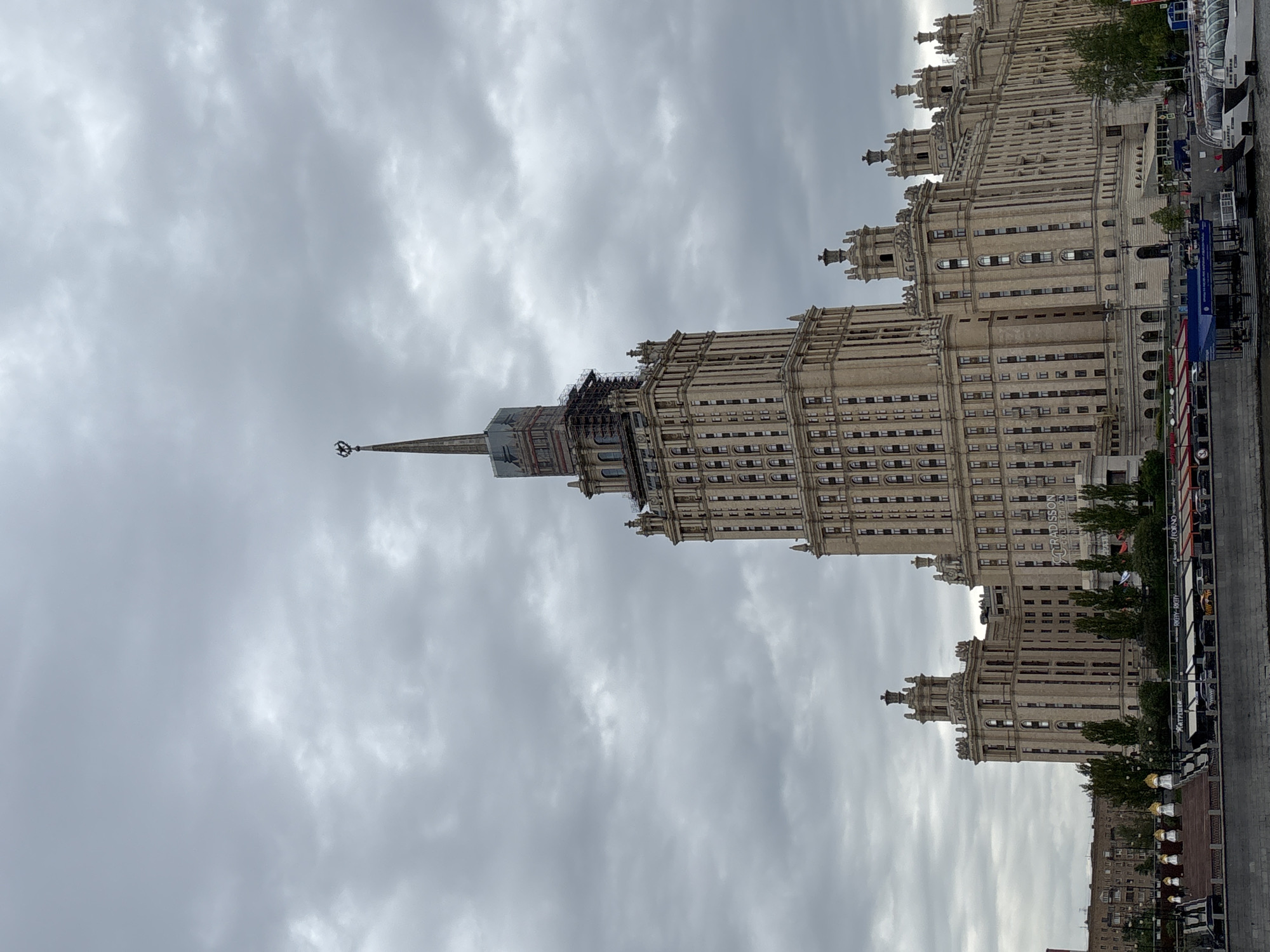 The image shows a tall, ornate building in Moscow, Russia, possibly one of the Seven Sisters skyscrapers. The building is constructed with a light-colored stone or concrete, featuring numerous windows and decorative architectural elements. It has a distinct spire topped with a star. The sky is overcast with gray clouds. Part of the city can be seen at the bottom of the image, including trees and some street furniture. The building appears to be undergoing some renovation or repair at the top of its spire.