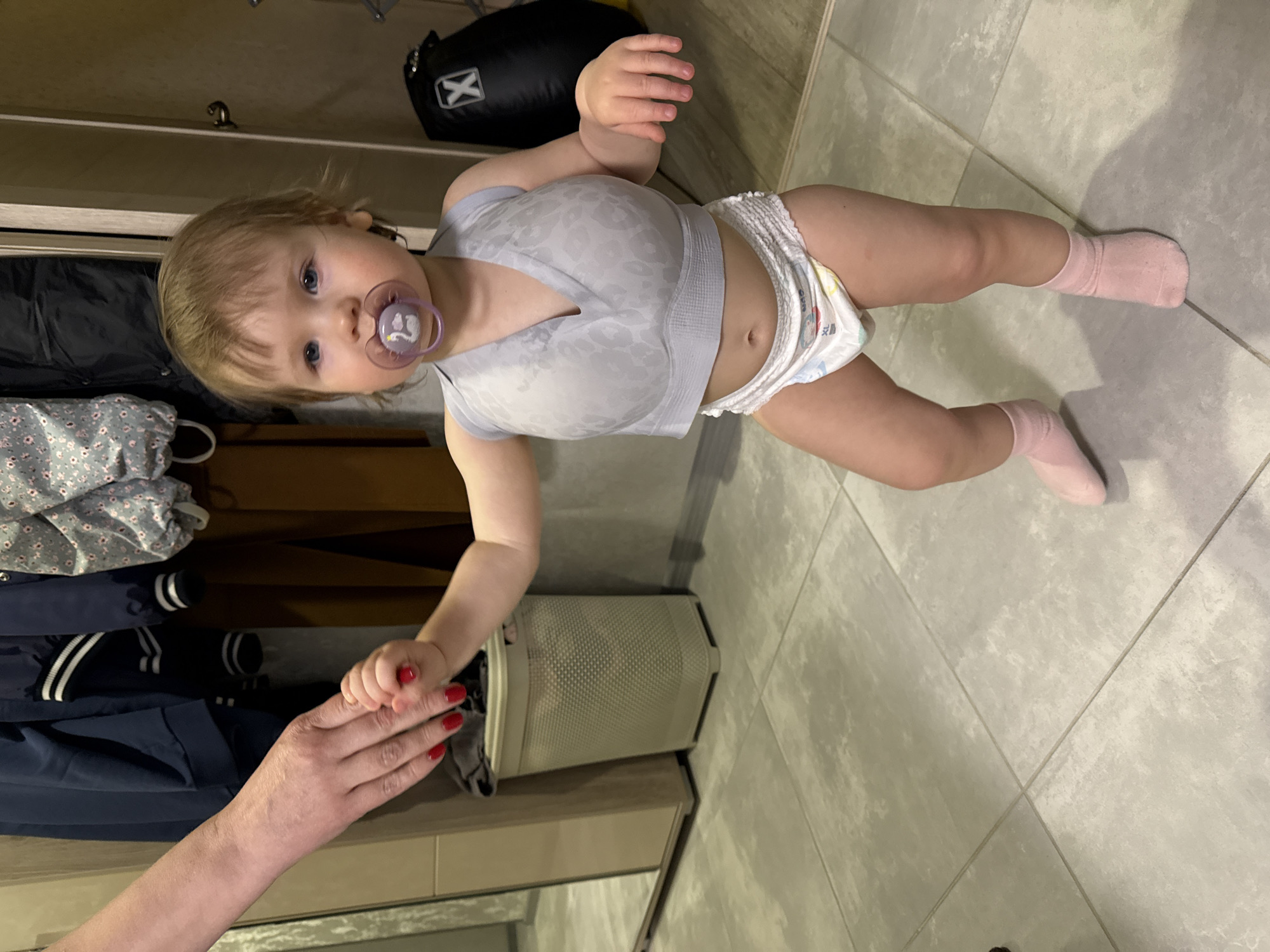 The image captures a top-down view of a baby or toddler standing indoors, holding an adult's hand. The baby has light brown hair and blue eyes, looking directly towards the camera with a neutral expression, and has a purple pacifier in their mouth. They are wearing a light grey V-neck top with a subtle leopard print pattern, a white disposable diaper, and pink socks.
The adult's hand, adorned with bright red nail polish, is extended to support the baby's small hand. The setting appears to be a home, with light grey tiled flooring visible throughout. In the background, there is shelving or a wardrobe containing dark clothing, including a navy jacket with white stripes. A black bag with a white "X" logo is placed on a shelf. To the baby's right, a light-colored, perforated bin or laundry basket is on the floor. The activity suggests the baby is being helped to stand or walk. The indoor lighting indicates either daytime or evening. The context for the location is Moscow, Russia.