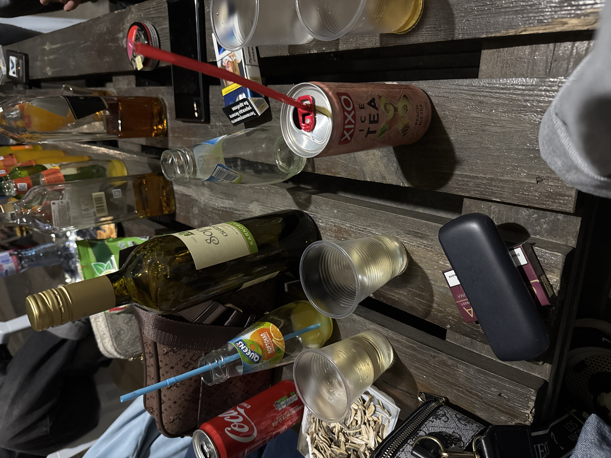 The image captures a seemingly casual social gathering, likely outdoors given the slatted wooden surface resembling a pallet or rustic table. Several beverages are present: a bottle of "SOP Chardonnay" wine, a can of "XIXO ICE TEA Peach," a "Coca-Cola" can, and an "QUEENS" juice drink with a straw. There are also plastic cups, some containing liquid, and several glass bottles, some empty. There is a dark glasses case and packs of cigarettes on the table. A purse and what appears to be a bag of sunflower seeds further contribute to the scene. It's difficult to precisely pinpoint a location in Topolovets, Bulgaria, with only these elements, but the presence of Bulgarian language on the cigarette pack and the "QUEENS" juice suggests a Balkan context. There are no people visible except part of a hand and part of a sleeve.