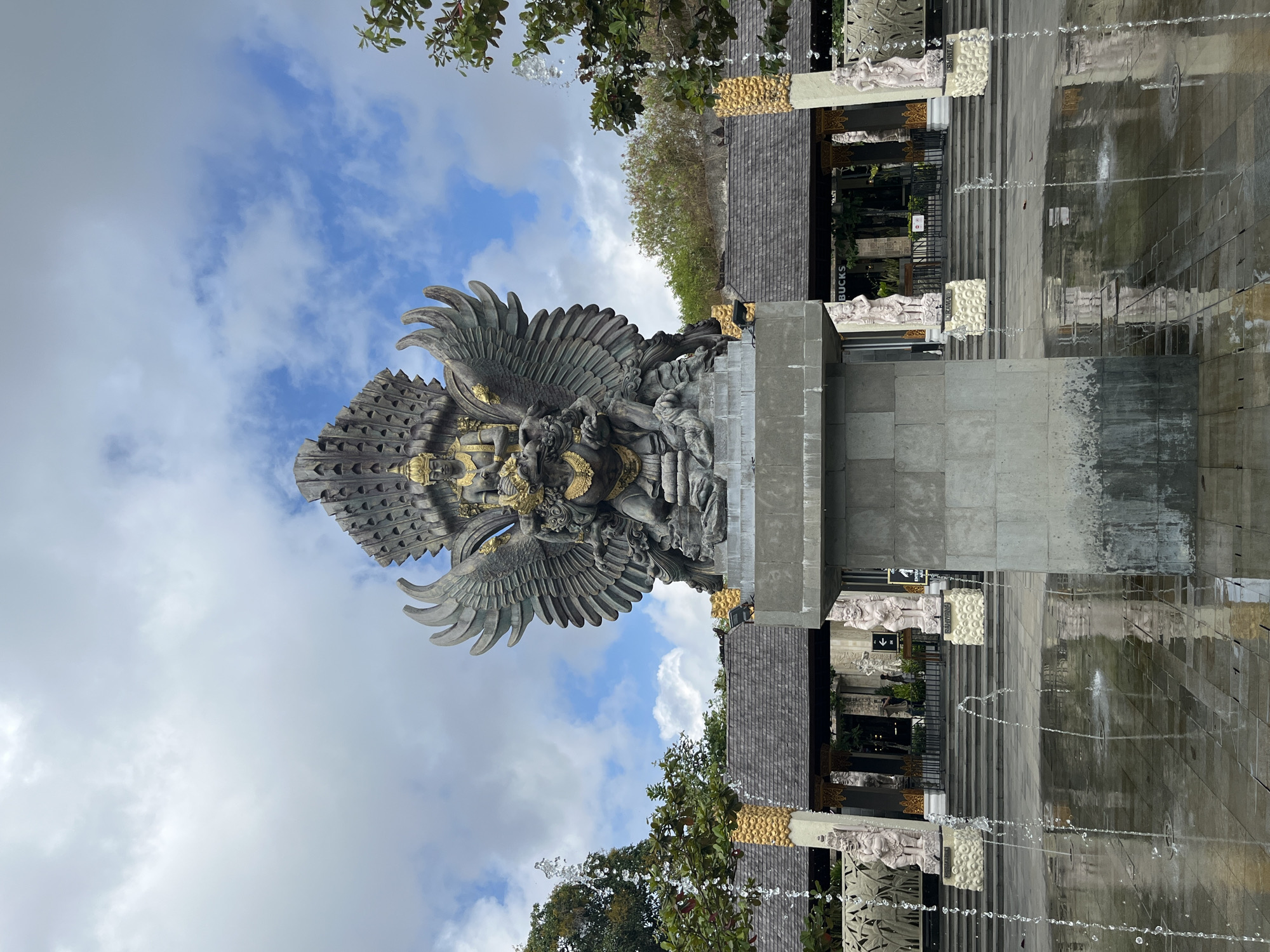 The image shows a large stone statue of Garuda, a mythical bird-like creature from Hindu mythology, in the city of Kuta Selatan, Indonesia. The statue is perched on a pedestal in front of a building with a traditional Balinese architectural style. The statue is intricately detailed, with Garuda depicted with outstretched wings and a human-like torso, seated upon a human-like figure. The background shows a cloudy sky and lush greenery. In the foreground, a fountain with several water jets creates a reflective surface on the paved ground, hinting at a public space or plaza. Several smaller stone statues of human-like figures are visible on either side of the building's entrance, flanking a set of stairs. A Starbucks sign is visible on the building, indicating a commercial establishment. The overall scene suggests a cultural or tourist attraction.