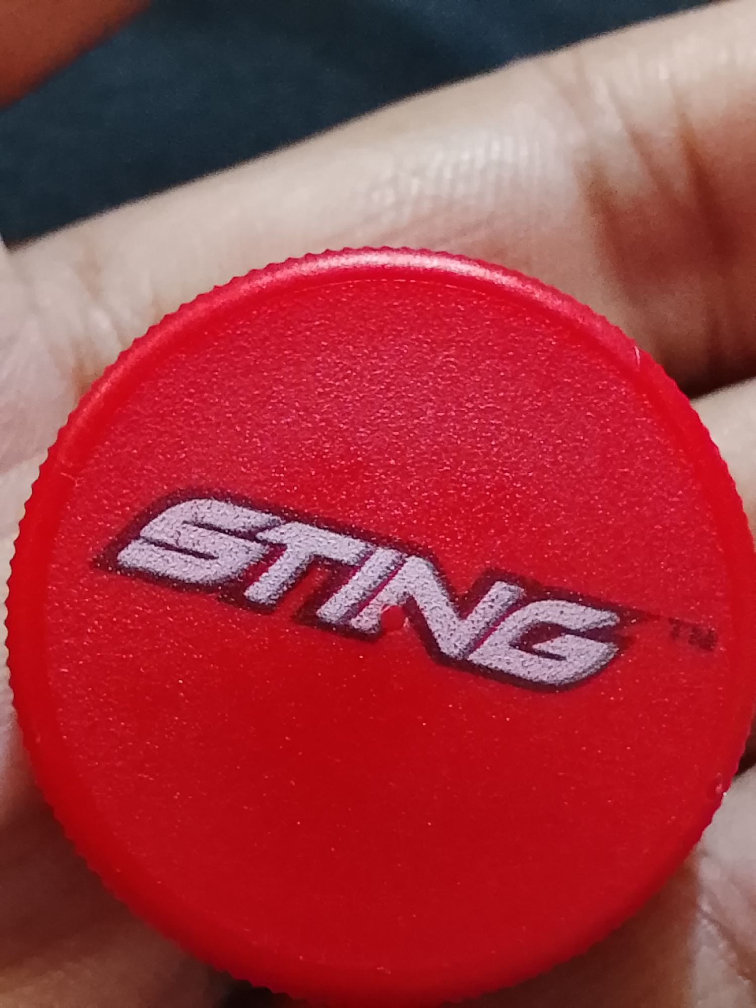 A red plastic bottle cap with the word "STING" printed on it in white with a darker outline. The cap has a textured edge. The image is close-up and slightly out of focus, showing a hand holding the cap. No information is available to determine the location in Multan, Pakistan.