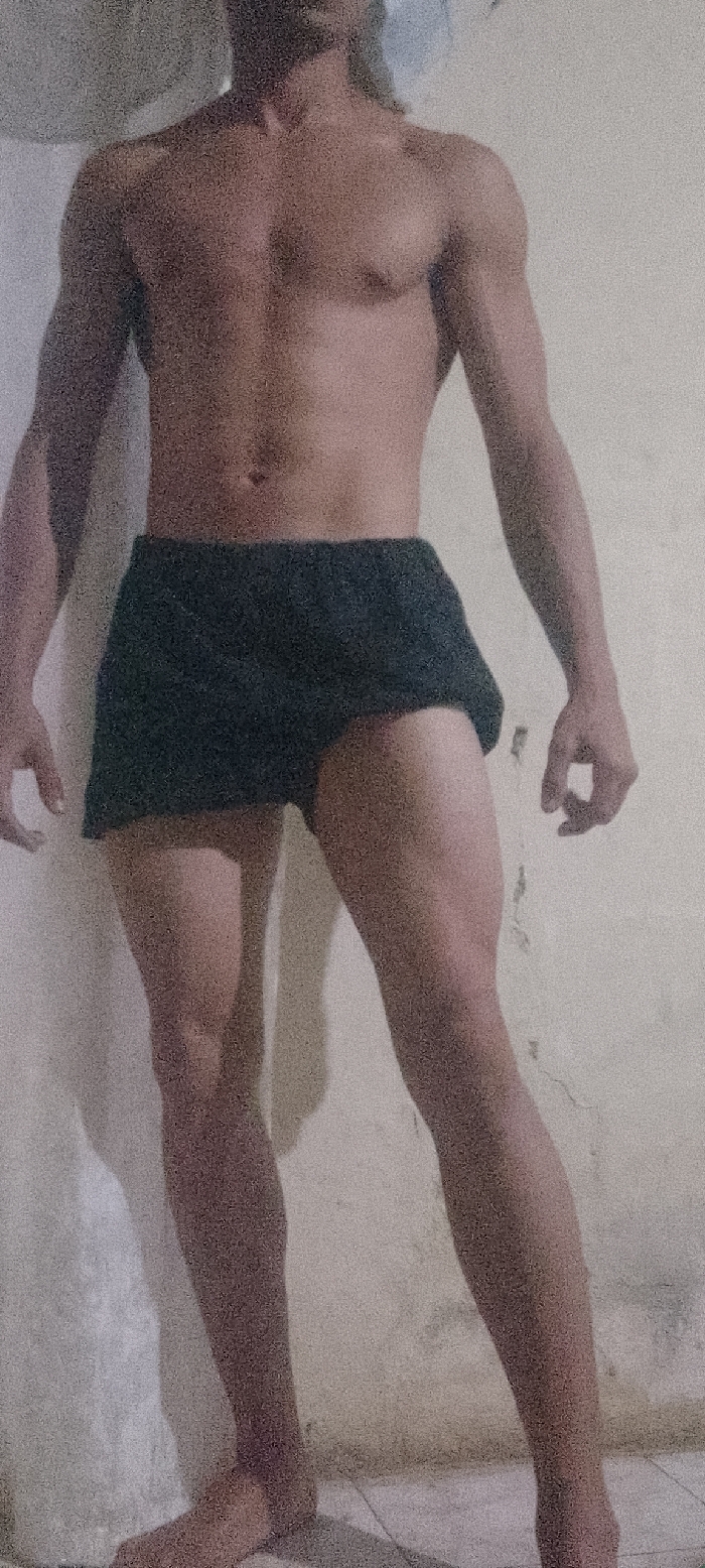 A full-body photograph captures an unidentified individual, presumed male, standing indoors in Juan Griego, Venezuela. The subject is shirtless, displaying a lean, muscular physique, and wears dark, possibly black, athletic shorts that extend to mid-thigh. The individual's head is cropped from the image, visible only from the lower neck and upper chest area. The subject stands with arms slightly bent and hands relaxed, with the left leg slightly forward and the right leg slightly back. The background consists of a light-colored, textured wall exhibiting minor imperfections and wear, including visible cracks and discoloration. The floor is covered with light-toned rectangular tiles with visible grout lines. Lighting is consistent, casting faint shadows to the left of the subject. The image exhibits significant digital noise, suggesting low light conditions or high ISO.