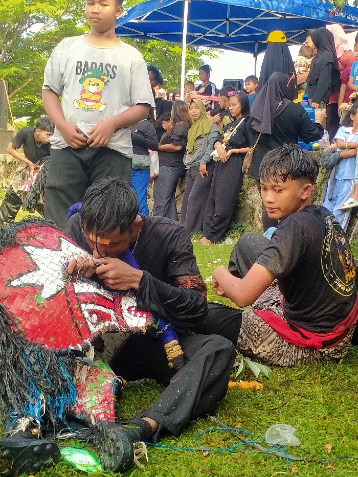 This image captures an outdoor gathering in Jetak, Indonesia. In the foreground, two young men are seated on green grass. The man on the left wears a black long-sleeved shirt and dark pants, has wet hair, and a small bandage on his forehead. He is intently working on a large, red, white, and green woven prop, possibly a mask or headpiece, adorned with black fibrous material. To his right, a second young man, also with wet hair, is seated, wearing a black short-sleeved t-shirt with a graphic emblem on the back and patterned lower garments with a red sash. He is looking towards the right side of the frame. Scattered on the grass are blue strings and plastic debris.
In the mid-ground, a young man stands upright, wearing a light grey t-shirt with a "BADASS" teddy bear graphic and black pants, observing the scene. Behind him, another person in a dark, shaggy costume is partially visible. Further back, under a blue canopy tent displaying "Bank Jateng" branding, a group of approximately a dozen adults and children are standing or leaning against a low concrete embankment. Notable figures include a woman in an olive-green hijab and another in a black hijab with a yellow baseball cap. A blue water dispenser and plastic cups are visible beneath the tent. The background reveals green trees and foliage under an open sky.