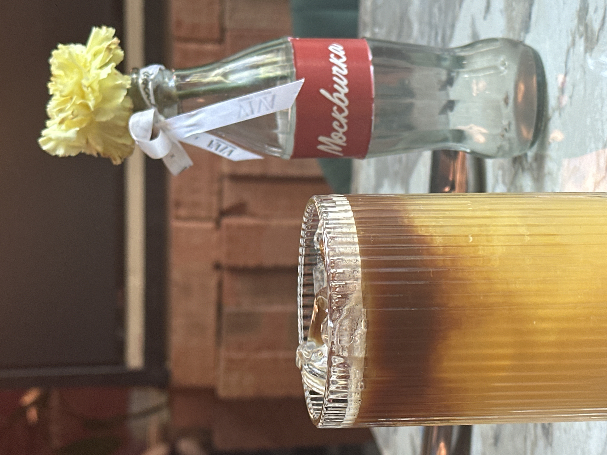 The image features a still life composition on a likely table or countertop. A clear glass bottle, labeled "Mockburka" in Cyrillic script on a red band, lies horizontally. A yellow flower is inserted into the bottle's opening, adorned with a white ribbon. Next to the bottle is a translucent, cylindrical glass container filled with a brown liquid. The background reveals a brick wall and a glimpse of what appears to be a window. The overall scene suggests a casual, possibly cafe setting, with a focus on simple decorative elements. It isn't possible to confirm the location in the city of Novosibirsk, Russia.
