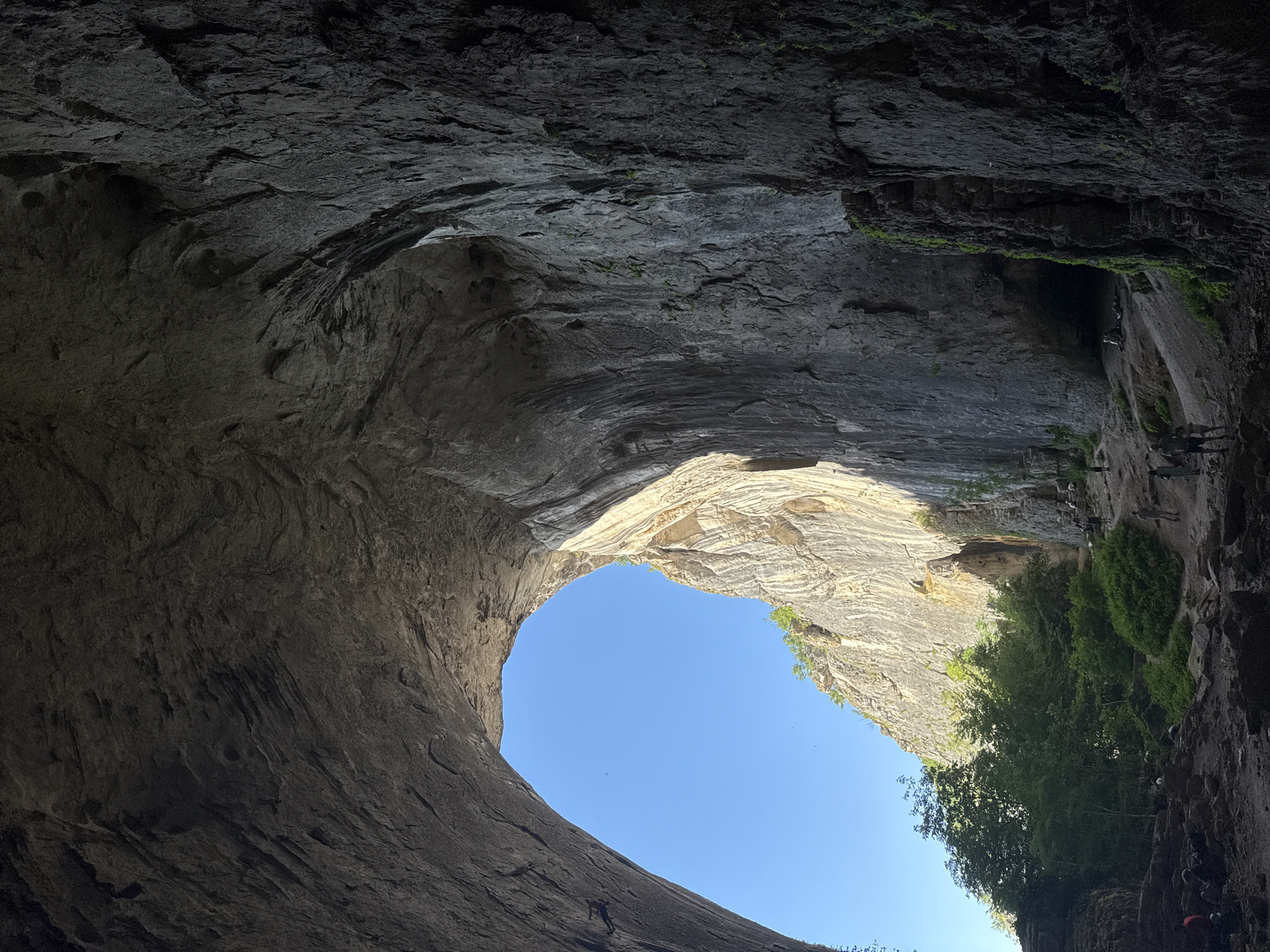 This image depicts the interior of the Prohodna Cave, also known as the "Eyes of God," located near the village of Karlukovo, Bulgaria. The cave is characterized by its massive scale, with towering rock formations forming a natural archway that frames a bright blue sky. Sunlight streams through this opening, illuminating the textured rock faces and casting shadows within the cavern.
The foreground of the image is dark and rocky, with uneven terrain suggesting a natural, unpaved surface. Various people are scattered throughout the scene, some appearing to be exploring or resting. In the mid-ground, a group of individuals is visible near the entrance of another section of the cave, with lush green foliage and trees growing against the cliff face. The rock walls themselves display a rough, layered texture, indicative of geological erosion over time. One rock face in the background shows a prominent, lighter-colored band, possibly a different mineral composition. The overall impression is one of a vast natural wonder, with human presence adding a sense of scale and exploration.