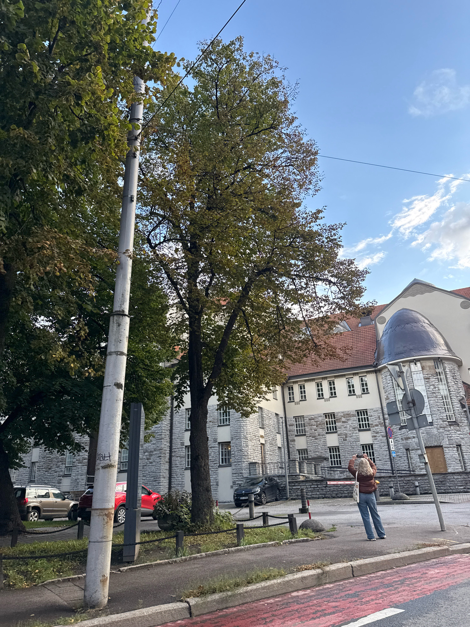 The image shows a woman standing on a sidewalk in front of a stone building with a domed roof in Tallinn, Estonia. She is holding a camera up to take a picture of the building. There are trees on the left side of the image, and a street with cars is visible behind her. The sky is blue with some clouds.