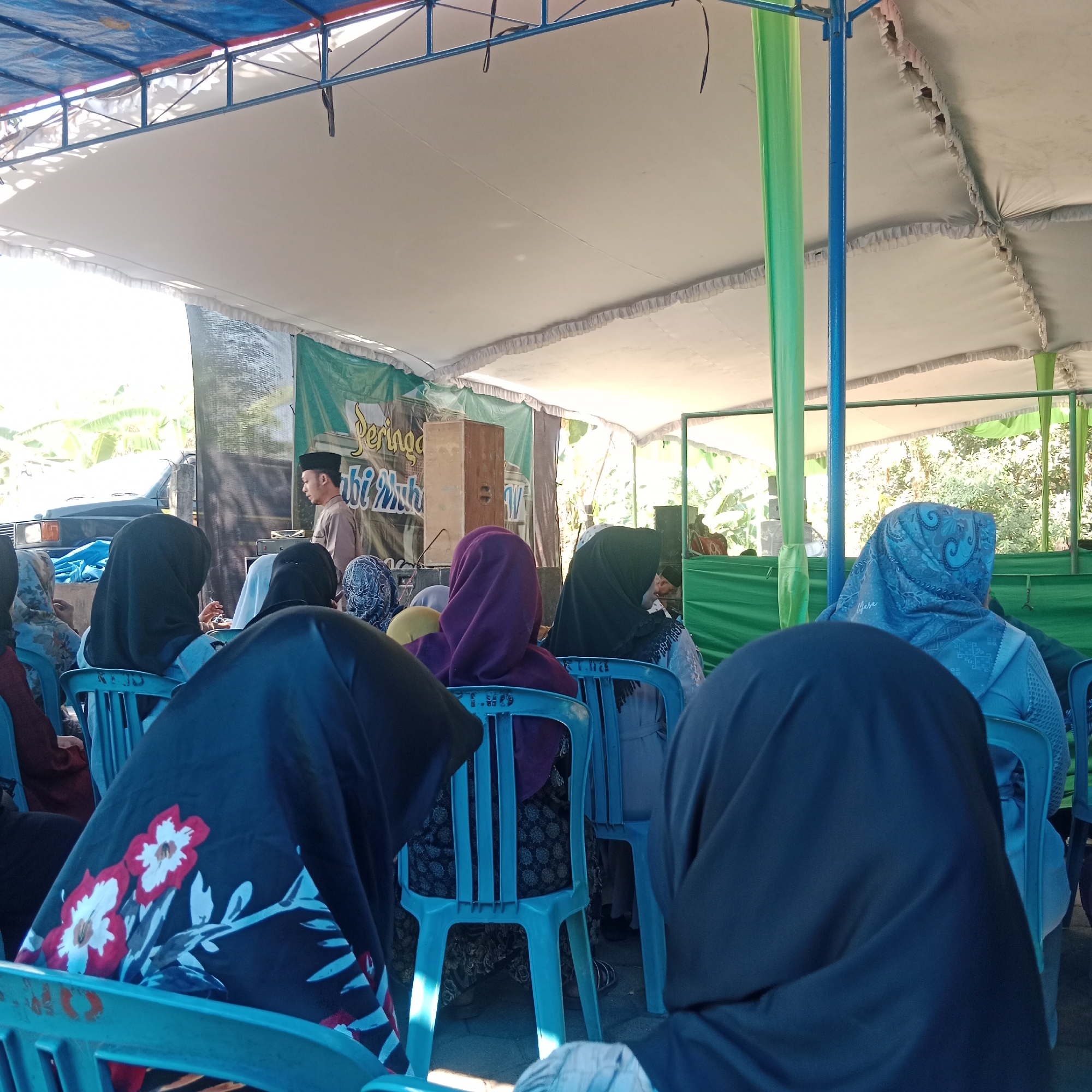 The image depicts an outdoor gathering in Tegowanu Kulon, Indonesia, likely a religious event or ceremony under a canopy. A man wearing a peci, a traditional brimless cap, and a light brown tunic stands near a wooden speaker box, addressing the audience. The audience, predominantly women dressed in hijabs of various colors and patterns including black, purple, blue, and grey, are seated on blue plastic chairs facing the speaker. A large banner with green and white lettering, partially obscured, is visible behind the speaker. The setting appears to be a community space, with trees and foliage visible in the background, suggesting a rural or semi-urban environment. The overall lighting indicates it is daytime, with sunlight filtering through the canopy.