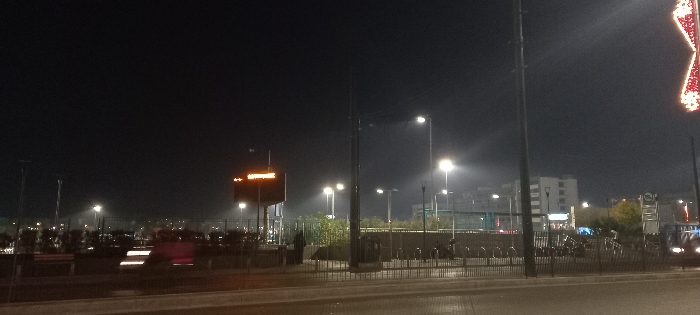 The image depicts an urban street scene at night in Piraeus, Greece. The sky is dark, illuminated by numerous artificial light sources, primarily bright streetlights that cast soft halos.
In the mid-ground on the left, a prominent rectangular orange sign glows, displaying white text. Closer inspection reveals the Greek word "ΑΦΕΤΗΡΙΑ" (meaning "STARTING POINT" or "DEPARTURE"), accompanied by a small arrow symbol pointing to the left. This likely indicates a bus stop or a departure point for transportation.
In the foreground, a dark road surface is visible. Along the edge of the road, a row of metal bicycle racks stands, some with bicycles parked in them. Behind the racks, a low fence separates the area from some sparse greenery. A tall, dark utility pole stands vertically in the center of the frame.
In the distance, several multi-story buildings with illuminated windows can be seen against the dark horizon, indicating a populated urban area. On the far right side of the frame, part of a large, brightly lit decorative red star is visible, possibly a festive ornament. On the road in the left-mid distance, blurred red and white light trails suggest the movement of vehicles. The overall scene is calm and showcases a typical nocturnal urban landscape.
