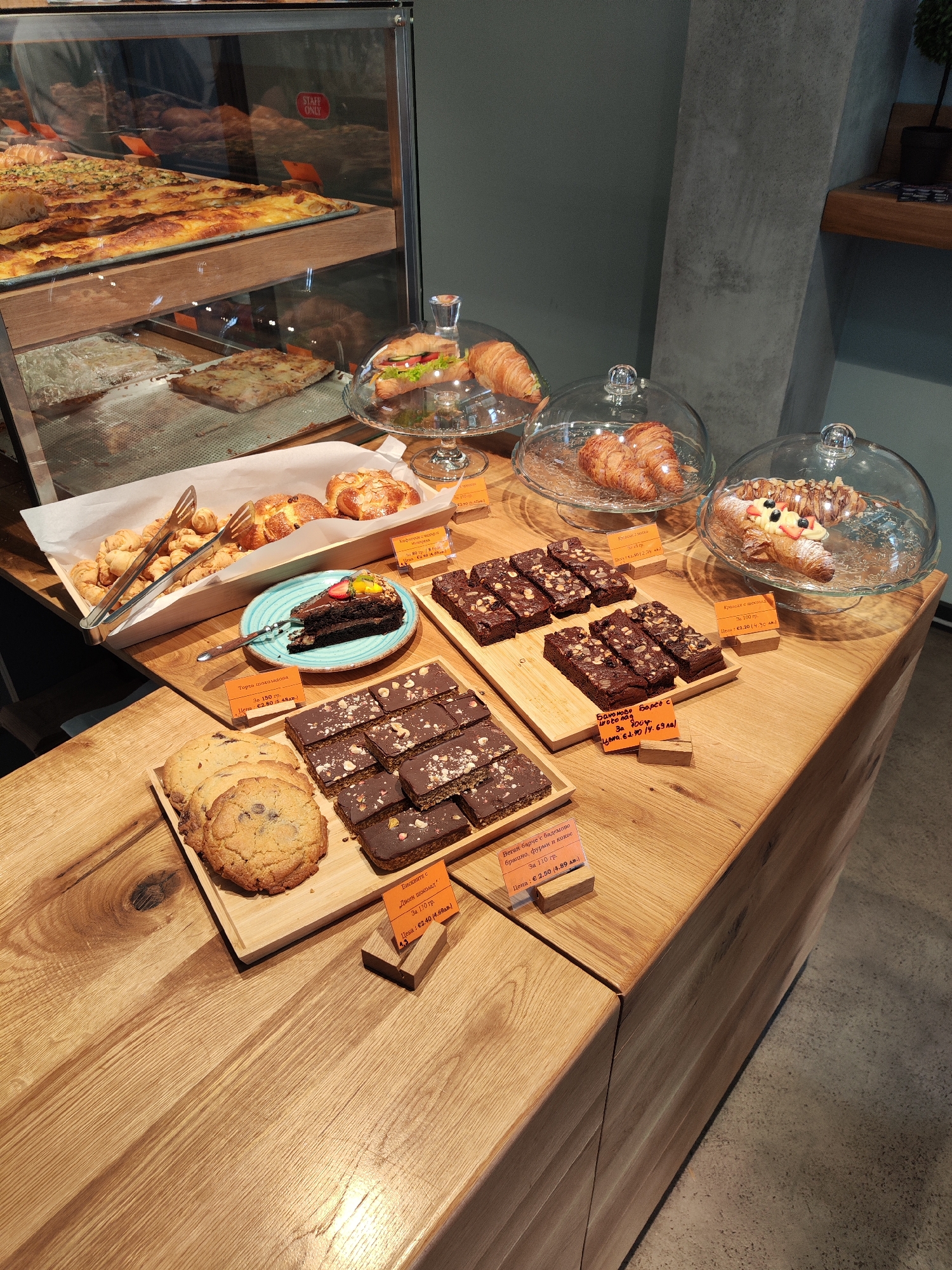 The image shows a bakery display with a variety of pastries and baked goods. On the top left, there is a glass display case with different types of pizza. In the center, there are trays of pastries including croissants, and a wooden tray with tongs, a slice of chocolate cake on a blue plate, and some small croissants.
To the right, there are more croissants under glass domes, along with several wooden boards displaying brownies and cookies, each accompanied by a price tag. The foreground features cookies and brownies, also with price tags.
The display is set on a wooden countertop. In the background, there is a grey wall.