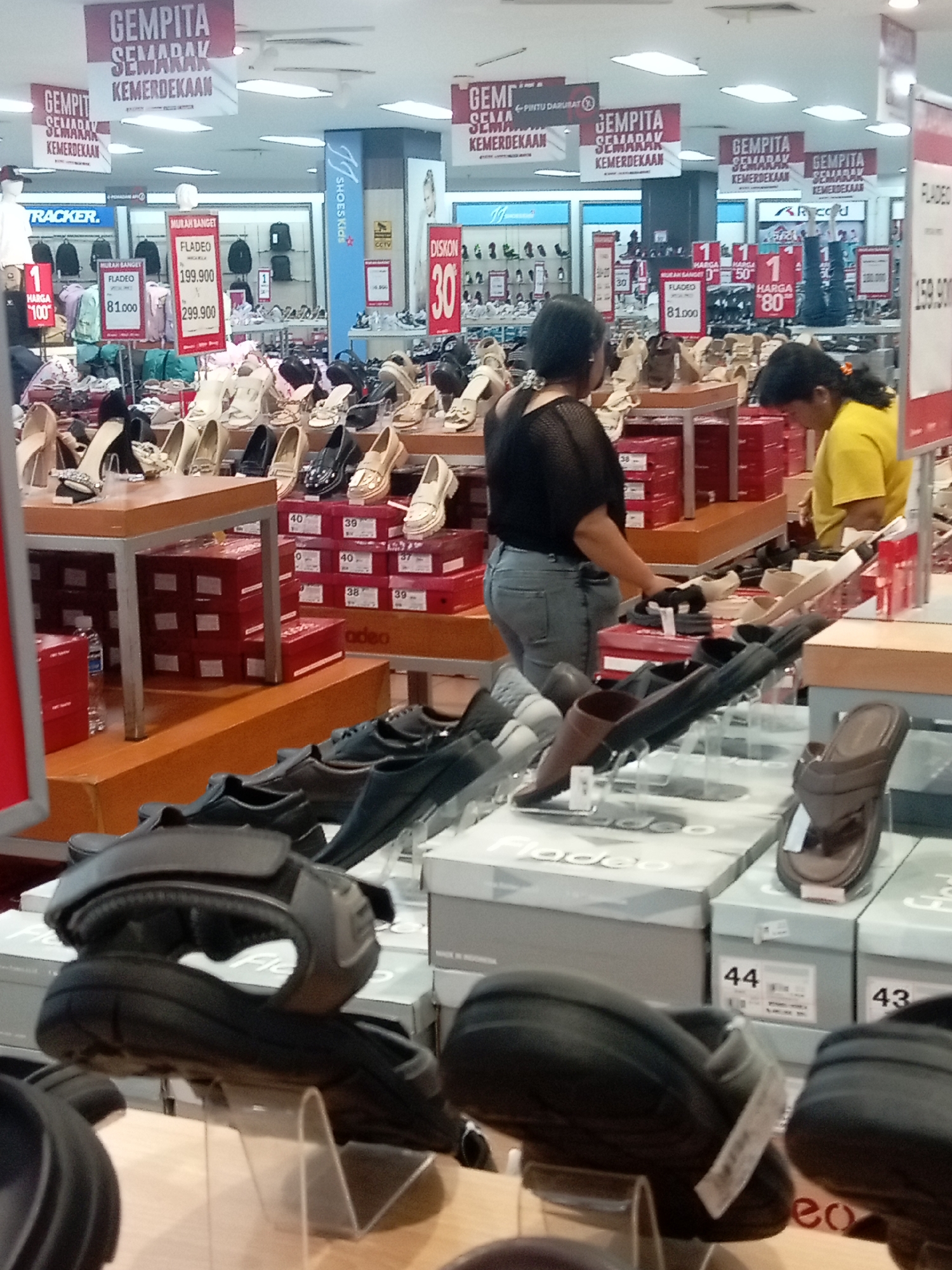 The image is a medium shot of a shoe store interior in Batam, Indonesia, featuring various footwear on display and sale. In the foreground, several pairs of women's sandals and heels are arranged on shelves and display stands, with a focus on comfort-oriented footwear like slides and chunky-soled sandals. In the mid-ground, more shoes are displayed on tiered tables, with shoeboxes stacked neatly below. Some of the shoeboxes have labels indicating sizes and brands. In the background, prominent red and white banners with Indonesian text, likely advertising a sale or promotion related to "Kemerdekaan" (Independence), are hung from the ceiling. Several shoppers are visible, including two women in the mid-ground, one examining shoes and another looking at a display. The environment appears to be a well-lit retail space, typical of a department store or a specialized shoe shop in a shopping mall.