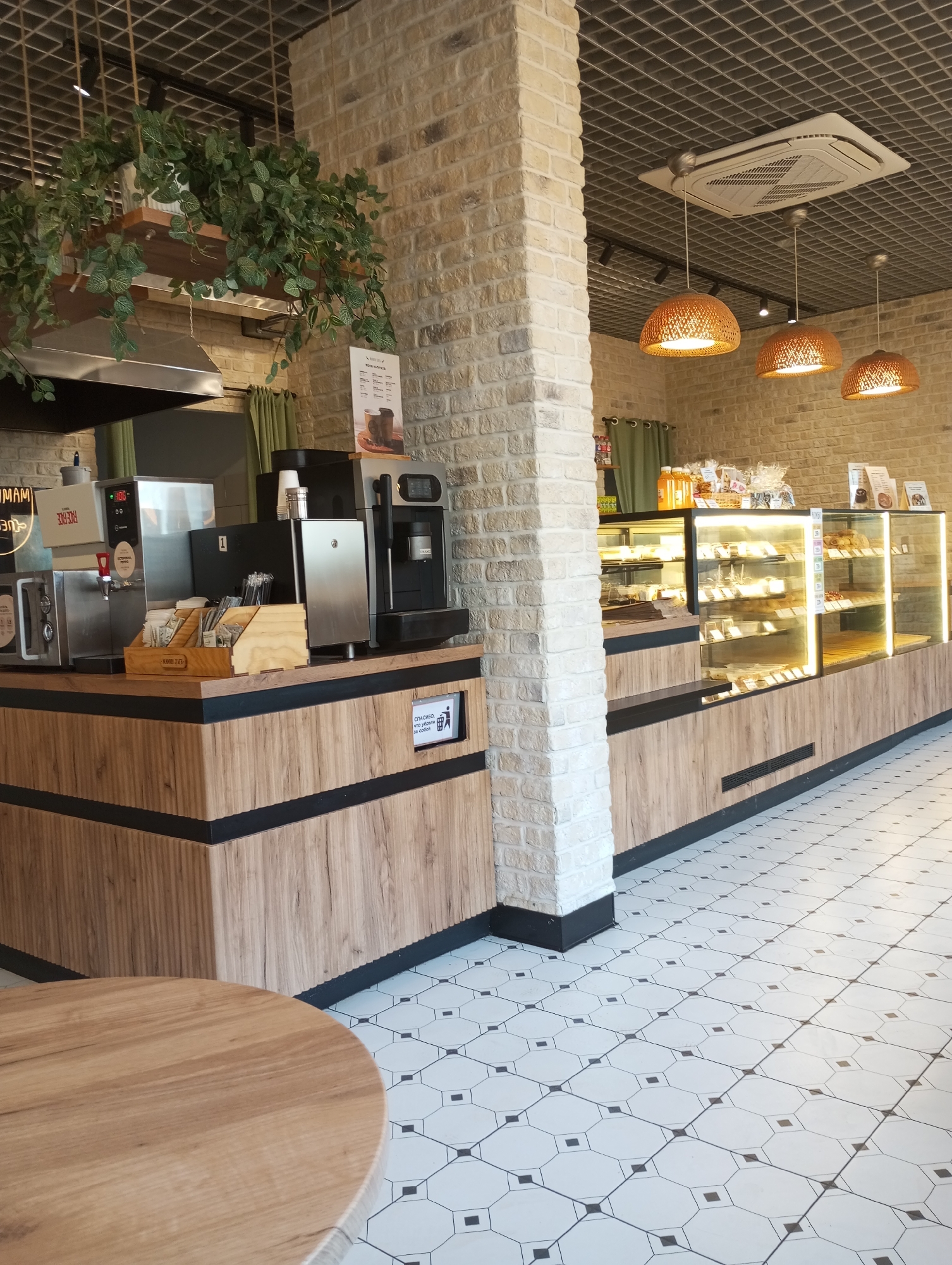 Here is a detailed description of the image:
**Scene Description:** The image captures the interior of a well-lit cafe or bakery setting. The decor includes brick walls, wooden counters, and a tiled floor with a geometric pattern. There's a service counter with a coffee machine and other drink preparation equipment. Display cases showcase baked goods. Potted plants hang above the counter area. Round light fixtures hang from the ceiling.
**Objects:**
* Coffee machine
* Display cases with baked goods
* Wooden counters
* Potted plants
* Tiled floor
* Round wooden table
* Light fixtures
**Environment:** The scene appears to be an interior space. The lighting is bright, and the design is modern with rustic elements.
**Location:** The OCR text shows that one of the objects in the picture reads KAMU ZAFE.
KAMU ZAFE is a cafe located in Pervomaysky, Russia. Thus, the image is taken in this city.