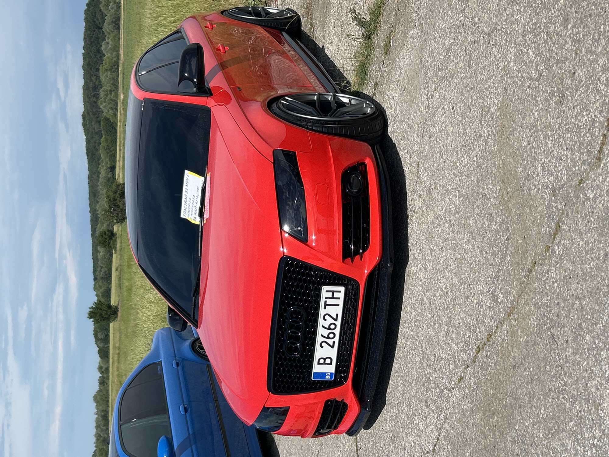 A close-up shot, oriented sideways, focuses on the front of a red car, likely an Audi, with a Bulgarian license plate reading "B 2662 TH" and a note placed on the windshield which reads, "ГЛАСУВАЙ ЗА МЕН С HOMEP 14 АНКЕТАТА ЗА НАЙ-КРАСИВ [ballot for me with number 14 the survey for the most beautiful]." The car is parked on a grey asphalt or concrete surface. In the background, a field of green grass and trees can be seen under a partly cloudy sky. Another blue car is partially visible on the left side of the frame. The scene is set in Dalbok Izvor, Bulgaria.