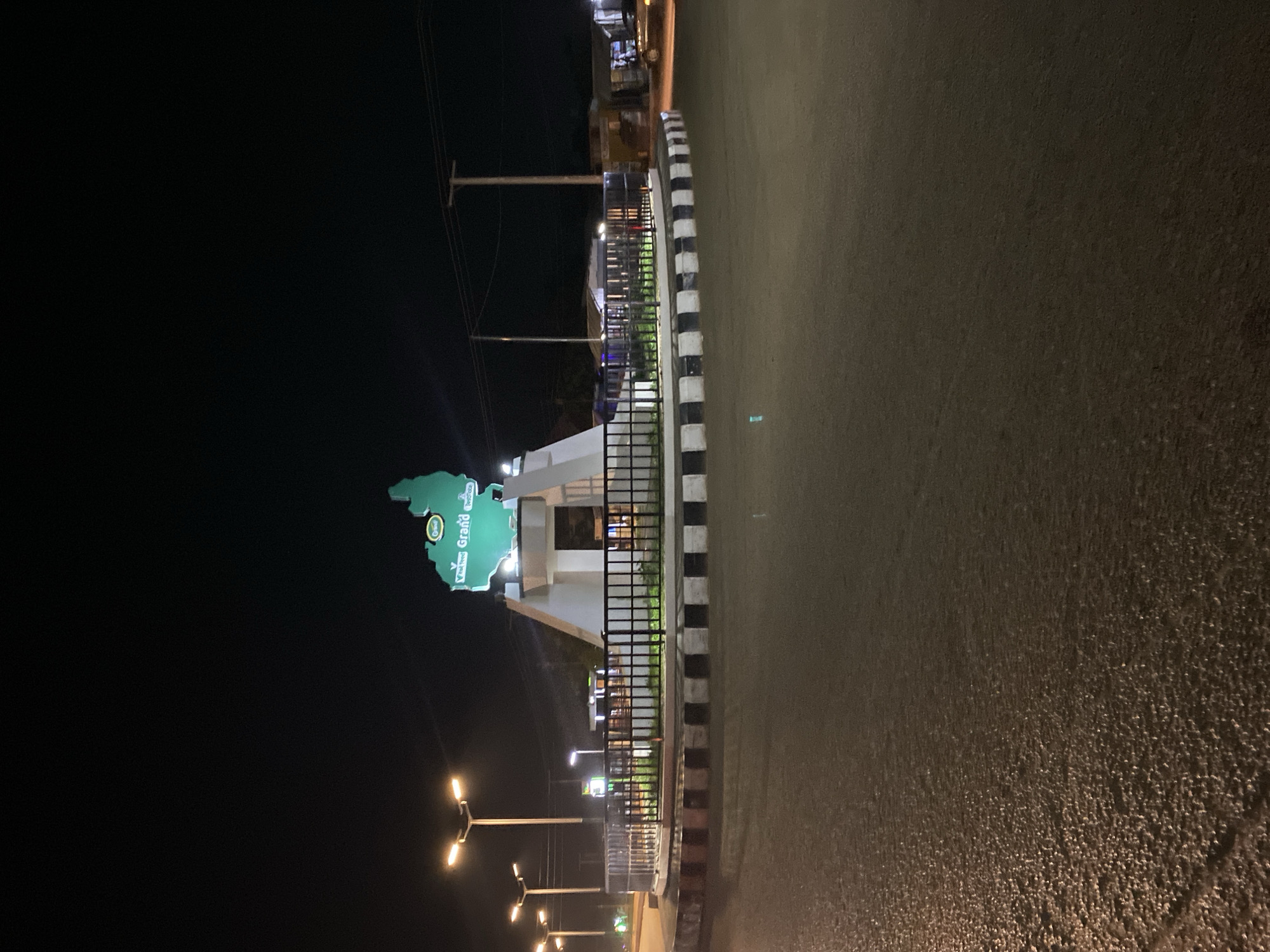 At night in Bukuru, Nigeria, a Yalkurum Grand Welcome sign is illuminated. The sign, shaped like a region of Nigeria, stands adjacent to a building with a black metal railing, which is behind a curb painted in black and white stripes. Street lamps provide additional illumination on the street. An electrical pole with wires is visible in the background. The paved road is wet.