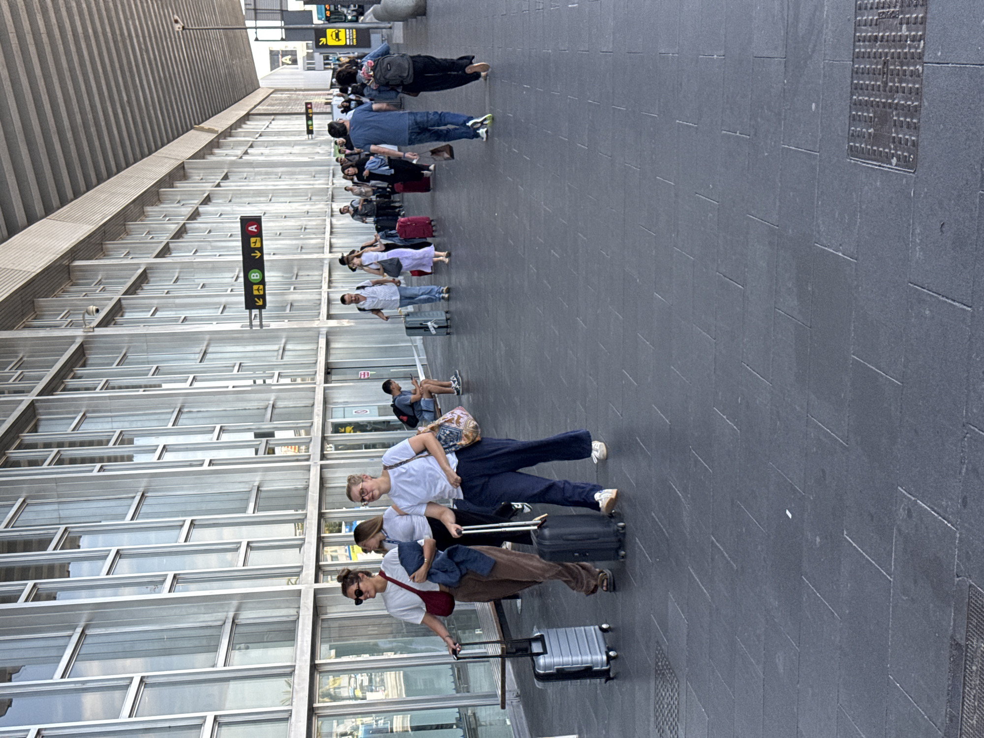 The image depicts a scene at Josep Tarradellas Barcelona–El Prat Airport in El Prat de Llobregat, Spain. A group of people are walking along an outdoor walkway in front of the airport terminal. The architecture features a modern facade with large glass windows and an overhead structure. In the foreground, two women are pulling suitcases. The woman on the left wears a white t-shirt, brown pants, and sunglasses, with a red bag slung over her shoulder. The woman on the right is dressed in a white t-shirt and dark pants, carrying a patterned tote bag. Further down the walkway, other travelers are visible, some with luggage, moving in the same direction. Signage with arrows and letters (A and B) is mounted on poles, indicating directions within the airport. The ground is paved with dark tiles. The overall atmosphere suggests a typical airport environment with people in transit.