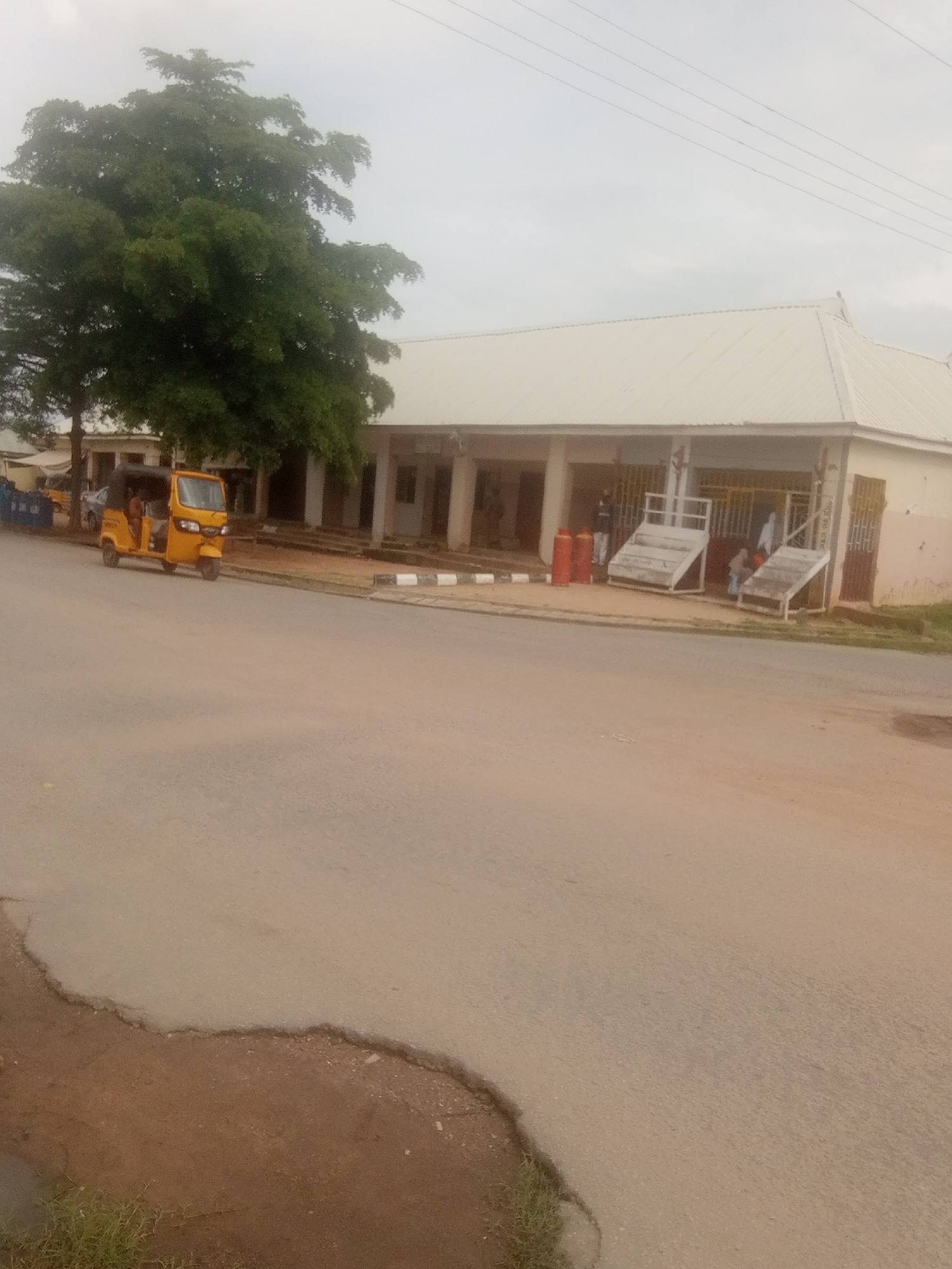 The image depicts a street scene in Jalingo, Nigeria, under an overcast sky, suggesting either early morning or a cloudy day. The primary focus is a yellow three-wheeled auto-rickshaw, commonly known as a "Keke Napep" or "Marwa," parked on the right side of the road. The Keke is empty and appears to be awaiting passengers.
In the background, a long, single-story building with a white corrugated metal roof stretches across the frame. The building has a covered walkway supported by pillars, and several windows or doorways are visible. Two people are standing under the covered area, and another individual is partially visible further down. In front of the building, there are two metal display stands or shelving units, possibly for goods. To the left of these stands, two large red gas cylinders are placed on the ground. A large, leafy tree with dark green foliage stands prominently to the left of the building, partially obscuring some of its structure.
The road in the foreground is paved but shows signs of wear and tear, with some cracks and uneven surfaces. The area to the left of the road is unpaved, with reddish-brown dirt and some sparse green vegetation. Overhead, several power lines are visible against the pale sky. The scene conveys a sense of everyday life in a Nigerian town, with commercial activity suggested by the building and the presence of gas cylinders. There is no discernible text in the image.