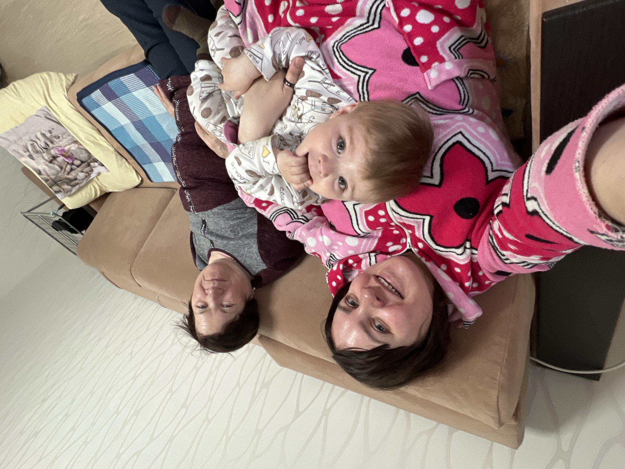 The image shows three individuals lying on a tan couch. A baby in a white onesie with cookies lies between two adult women. The woman on the right is wearing a pink blanket. The woman on the left is wearing a gray and burgundy top. A blue and white blanket, a yellow pillow, and dark clothing are also visible on the couch. The image was taken indoors, the background features white wallpaper and a dark-colored piece of furniture. The location is the city of Perm, Russia.