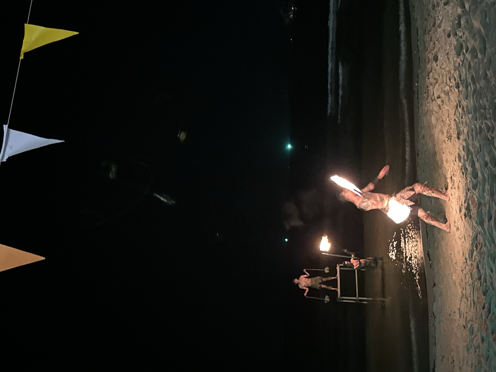 Night scene on a beach in Baan Klong Prao, Thailand. Three triangular flags—yellow, white, and orange—are strung along a line in the upper left corner. Two people are performing with fire on the beach. One person is near the water's edge with two flaming torches mounted on a stand. The other person, covered in tattoos, is lying on the sand with two torches and one in their mouth. Light from the flames reflects off of the wet sand near the water.